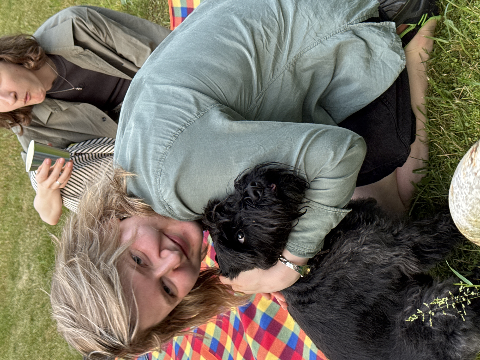 This is a photograph taken outdoors on a grassy area. The foreground is dominated by a person embracing a black Schnauzer dog. The person has blonde hair and is wearing a green shirt. They are looking towards the camera with a smile. The dog is black with a shaggy coat and is being held by the person. In the background, another person is visible, holding a silver cup and looking away from the camera. They are wearing a khaki-colored jacket over a dark shirt. The ground is covered in green grass and a colorful checkered blanket. The lighting suggests it is daytime. There are no identifiable Moscow landmarks or indicators of its location within the city.