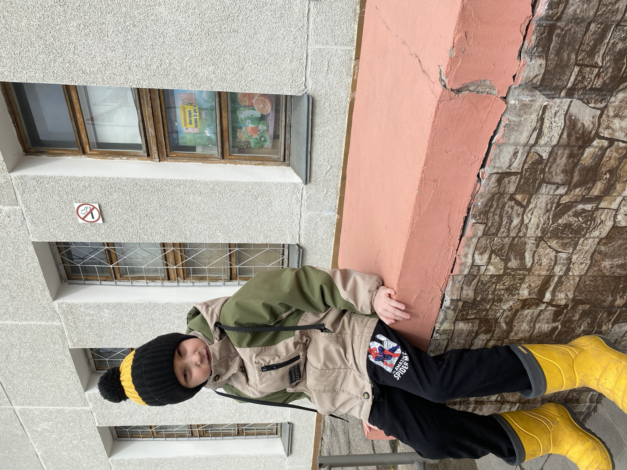 A child wearing a black and yellow beanie, a beige and green jacket, black pants with a Spider-Man graphic, and yellow boots stands outdoors. The child is next to a building with gray stucco walls, multiple windows, a pink corner column with cracks, and a decorative brick wall pattern. The location is the city of Chaikovsky, Russia.