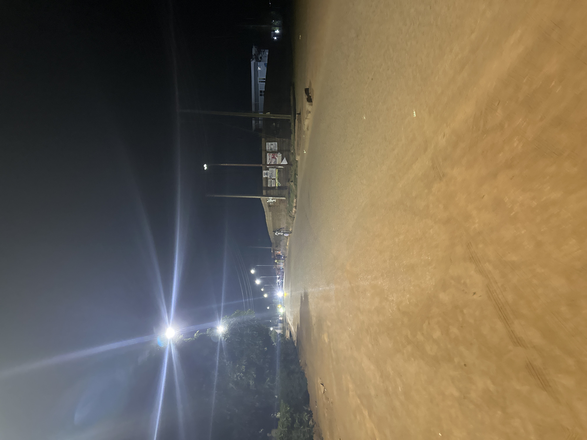 The image shows a street scene at night in Bukuru, Nigeria. The street stretches into the distance, illuminated by streetlights. Utility poles line the left side of the road, and a building with lit windows is visible in the background. Tire tracks are visible on the pavement. Some trees are visible to the left.
