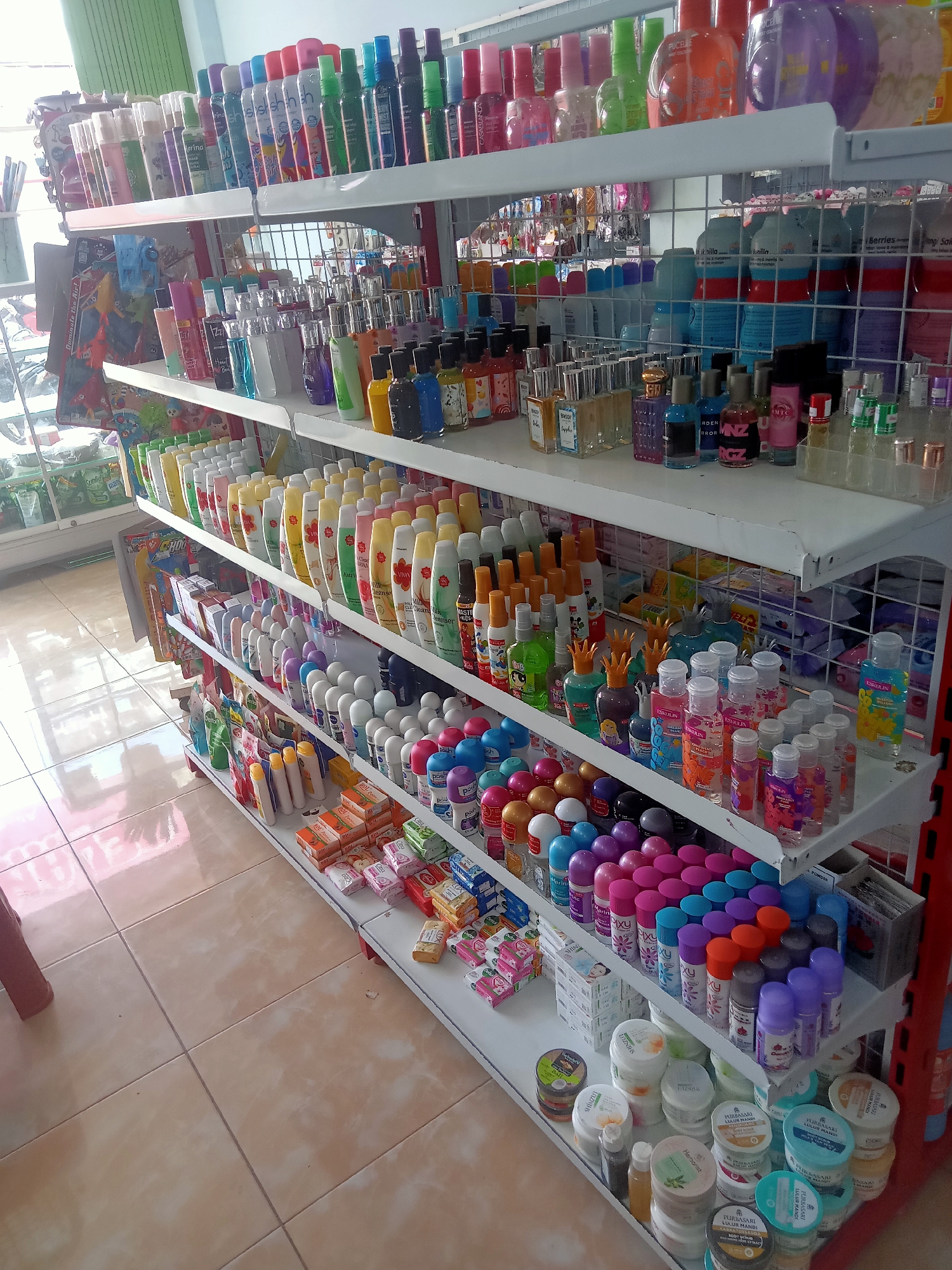 The image shows a retail store, likely a minimarket or convenience store, in Pule, Indonesia. The store shelves are stocked with a variety of personal care products, including lotions, perfumes, body sprays, deodorants, and soaps. The products are predominantly bottled and packaged in a range of colors and sizes, suggesting a diverse inventory catering to various consumer needs. The shelves are organized and well-lit, with products neatly arranged for display. The floor is tiled in a neutral, light brown color. The store appears to be tidy and well-maintained, with a clean and organized presentation of merchandise. The overall impression is that of a typical small-scale retail establishment focused on everyday consumer goods.