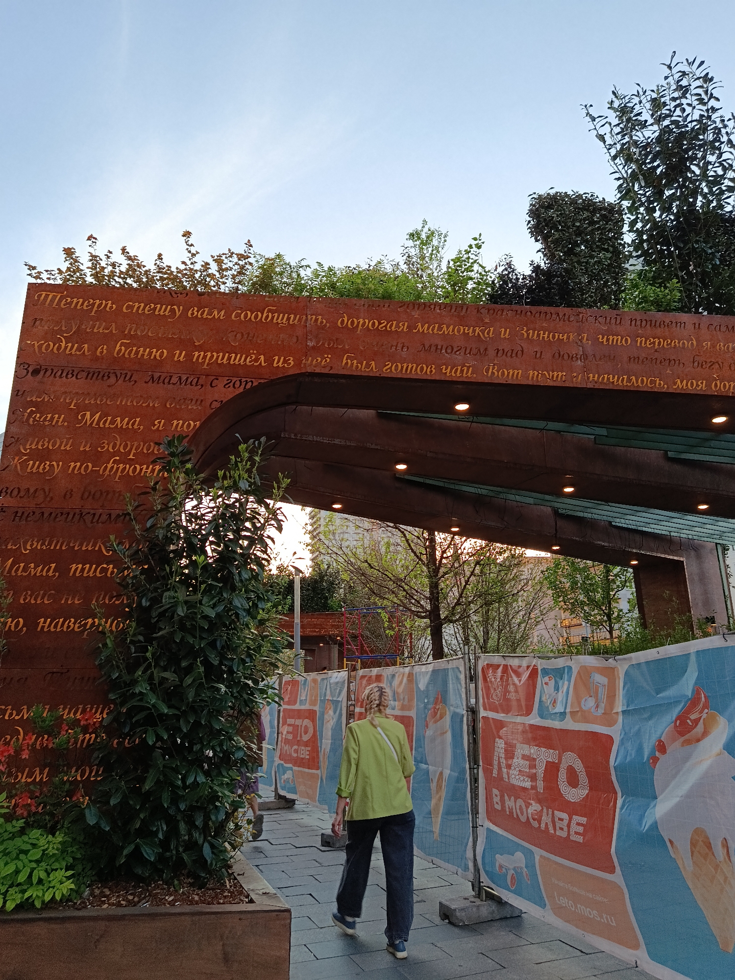 The image shows an outdoor scene in Moscow, Russia. A woman in a green shirt and dark pants is walking away from the camera on a paved path. To her right is a fence with banners advertising "Summer in Moscow" ("Лето в Москве"). Behind the fence, there is a park area with playground equipment visible.
On the left, there is a tall, rusted metal structure resembling an archway. The structure is covered in Russian text written in a golden color. Bushes and small trees grow on top and around the structure. The overall atmosphere is of a public park or pedestrian area undergoing some form of construction or renovation.