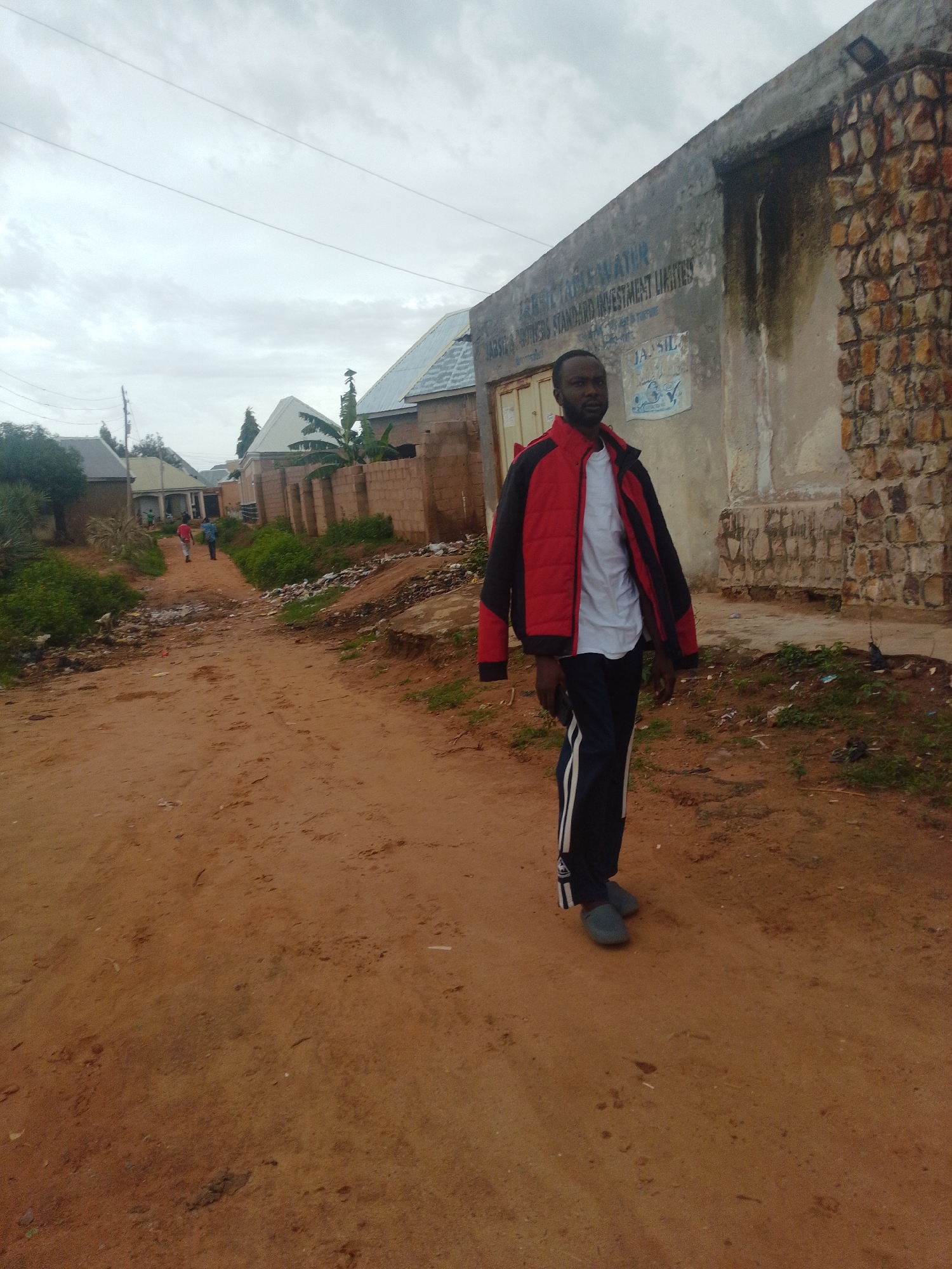 A man in a red and black jacket, white t-shirt, and dark pants with white stripes is walking on a dirt road in Tempure, Nigeria. The road is reddish-brown and appears to be in a rural or semi-urban area. To the right of the road is a building with a stone facade and a weathered, possibly concrete, wall with some dark stains. Text is visible on the building's wall, including "TABLE WATER" and "INVESTMENT LIMITED". There are some trees and other buildings in the background, as well as a pile of trash on the right side of the road. The sky is overcast. Two other people, one in a red shirt and one in a blue shirt, are visible walking away from the camera in the distance on the dirt road. The scene appears to be a street in a Nigerian town, with a mixture of residential and commercial structures.