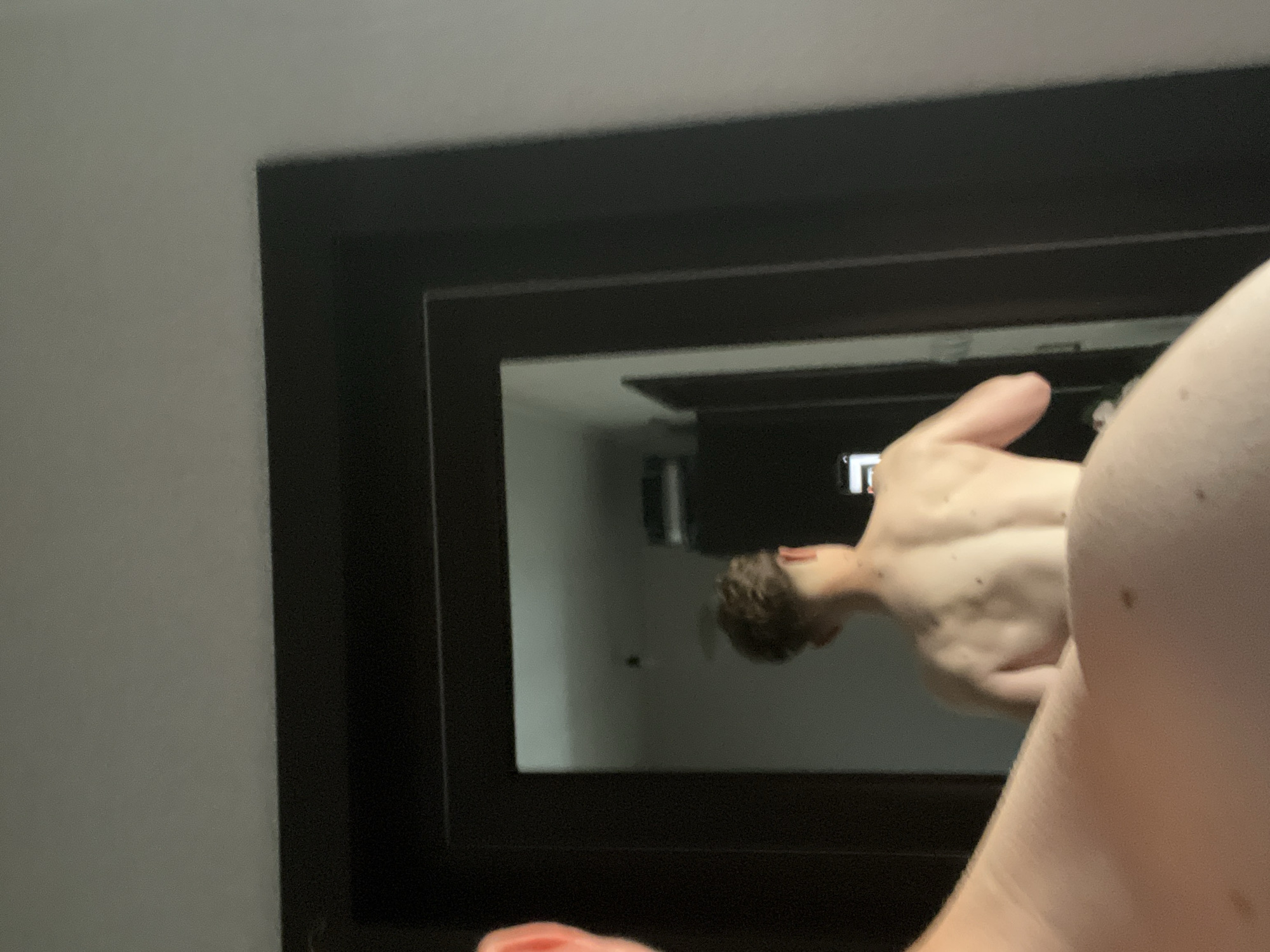 The image shows a person's reflection in a black-framed mirror. The person has fair skin and short brown hair. Part of their torso and arm are visible in the foreground, with moles on their shoulder. The reflection shows the person partially turned, with their hand reaching forward. The background is a blurry interior space, possibly a room. The location is not discernable from the image.