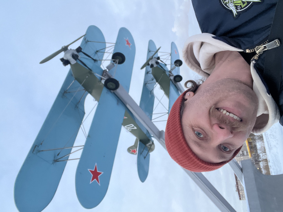 The image is a selfie featuring a smiling man in the foreground, with several biplanes displayed outdoors in the background. The photograph was originally taken in a portrait orientation but is rotated 90 degrees clockwise in the provided file.
The man, who occupies the right side of the frame (when viewed in correct orientation), has fair skin, blue eyes, and a light brown mustache. His hair is curly and light brown, visible around his ears. He is wearing a reddish-orange knit beanie, a dark blue jacket, and a light-colored (possibly white or off-white) hooded sweatshirt underneath. A metallic, rectangular zipper pull with a textured, grid-like pattern is prominently visible on his jacket. A patch depicting green figures or symbols on a dark background is visible on his right shoulder. He is looking directly at the camera with a wide, cheerful smile, showing his teeth.
In the mid-ground and background, at least three biplanes are arranged vertically, as if in the process of taking off or in an upward flight attitude. The planes have light blue wings and fuselages that appear to be a muted olive green or khaki color. Each biplane displays a distinctive red star insignia on the underside of its lower wing. One of the planes clearly shows the number "20" painted in white on its fuselage. Propellers are visible on the front of each aircraft, and small black landing gear wheels are attached underneath the lower wings. The biplanes appear to be part of an outdoor monument or museum exhibit.
The sky in the background is bright and overcast, suggesting a cloudy day. Faint outlines of distant buildings or structures are visible on the horizon, and some light-colored ground or structures are present beneath the planes, possibly indicating snow or ice on the ground, hinting at a cold environment.