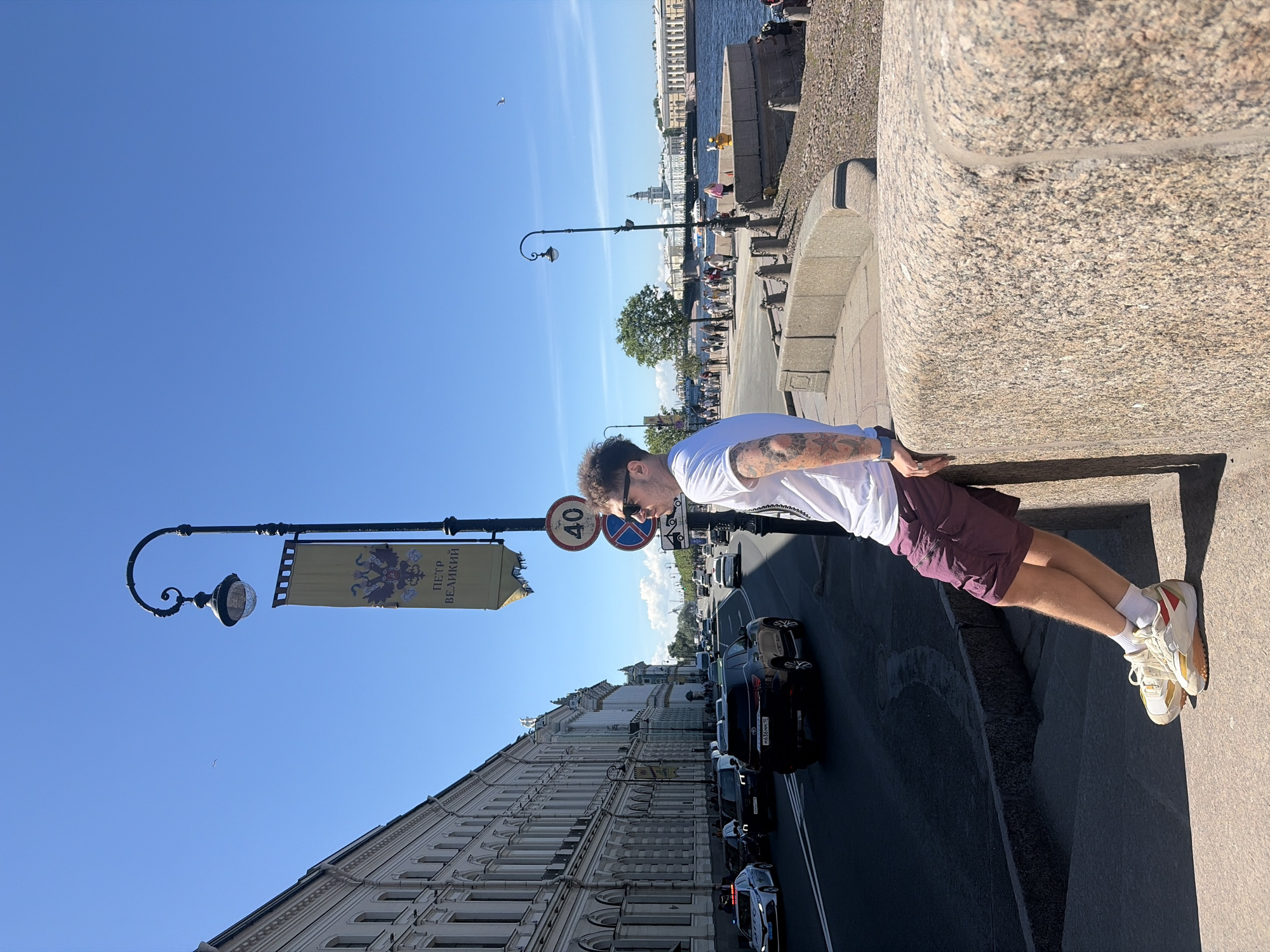 The image captures a sunny, clear day on the Palace Embankment in St. Petersburg, Russia, looking across the Neva River towards Vasilyevsky Island. In the foreground, a man in his late twenties or early thirties is leaning against a textured, light-brown granite parapet, facing right and looking slightly upward and away from the camera. He has short, curly dark hair, is wearing dark sunglasses, a white short-sleeved t-shirt, maroon cargo shorts, white socks, and off-white sneakers with yellow and red accents. Tattoos, including a star design, are visible on his left arm.
To the left of the man, a busy multi-lane road is visible with several cars, including a black SUV with a Russian license plate (H836MM 178) and a white sedan, possibly a police or emergency vehicle, with a blue light bar. Alongside the road, a grand, light-colored building in a classical architectural style stretches into the distance, featuring numerous arched windows and decorative elements. A black ornate lamppost stands prominently, bearing a yellow banner with a double-headed eagle emblem and the Cyrillic text "ПЕТР ВЕЛИКИЙ" (Peter the Great). Below the banner, two circular road signs are visible: a "40" speed limit sign and a "No Stopping" sign.
To the right of the man, beyond the parapet, the wide, dark blue Neva River flows. A stone-paved promenade runs parallel to the river, with several pedestrians walking. Chain barriers are visible along the river's edge. A small, leafy green tree stands between the promenade and the river. In the midground across the river, more grand, light-colored buildings with classical architecture line the distant bank, including a distinctive building with a prominent domed tower, likely the Kunstkamera or a similar structure on Vasilyevsky Island. The sky above is a clear, bright blue with a few wispy white clouds, and a small white bird is visible in flight in the upper left quadrant. Sunlight casts sharp shadows, indicating a high sun position.