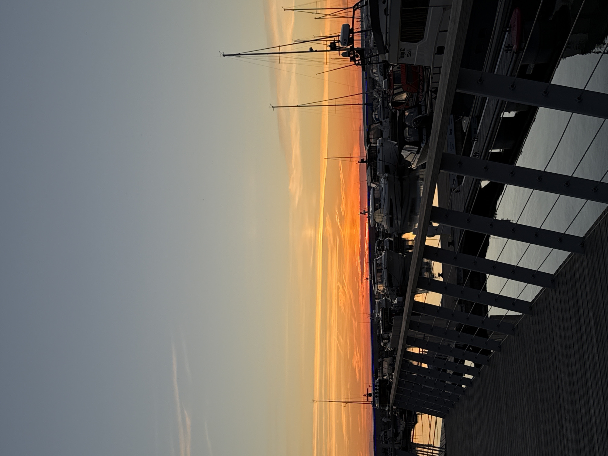 The image captures a sunset over a harbor in Tallinn, Estonia. A wooden pier with a metal railing extends into the foreground, leading towards a body of water where numerous boats are docked. The sky displays a gradient of colors, with a bright orange glow near the horizon transitioning to a pale blue at the top. The water reflects the colors of the sky and the silhouettes of the boats. The boats are of various sizes and types, including sailboats and motorboats. The scene is tranquil and serene, indicative of the end of the day.