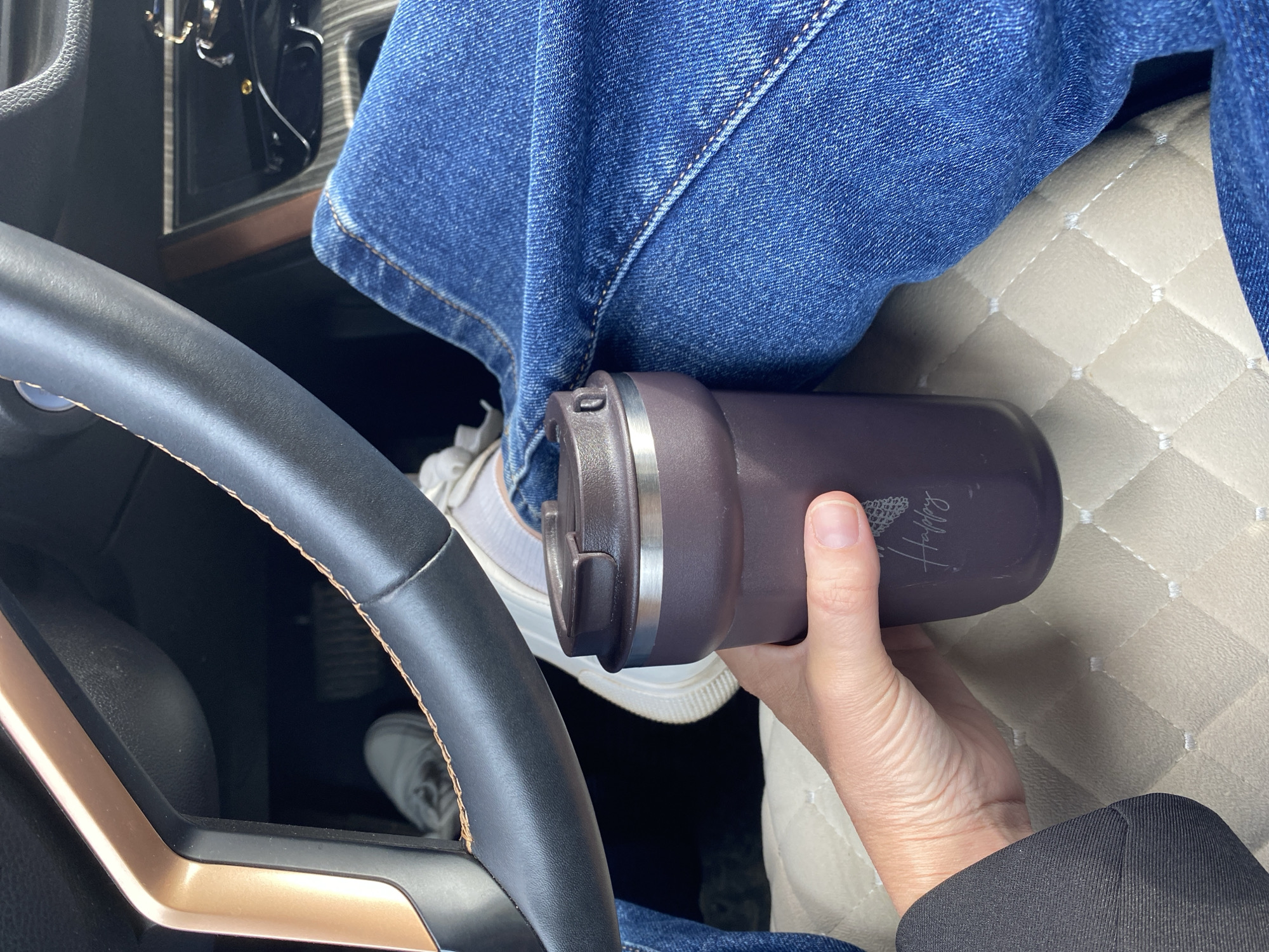 A person wearing blue jeans and a black blazer is seated in a car, holding a dark purple tumbler with the word "Happy" inscribed on it. The tumbler sits atop a white sneaker. A black steering wheel with a gold-colored accent is visible to the left, while a quilted beige surface is seen on the right. The specific location within the city of Chaikovsky, Russia is not identifiable from this image.