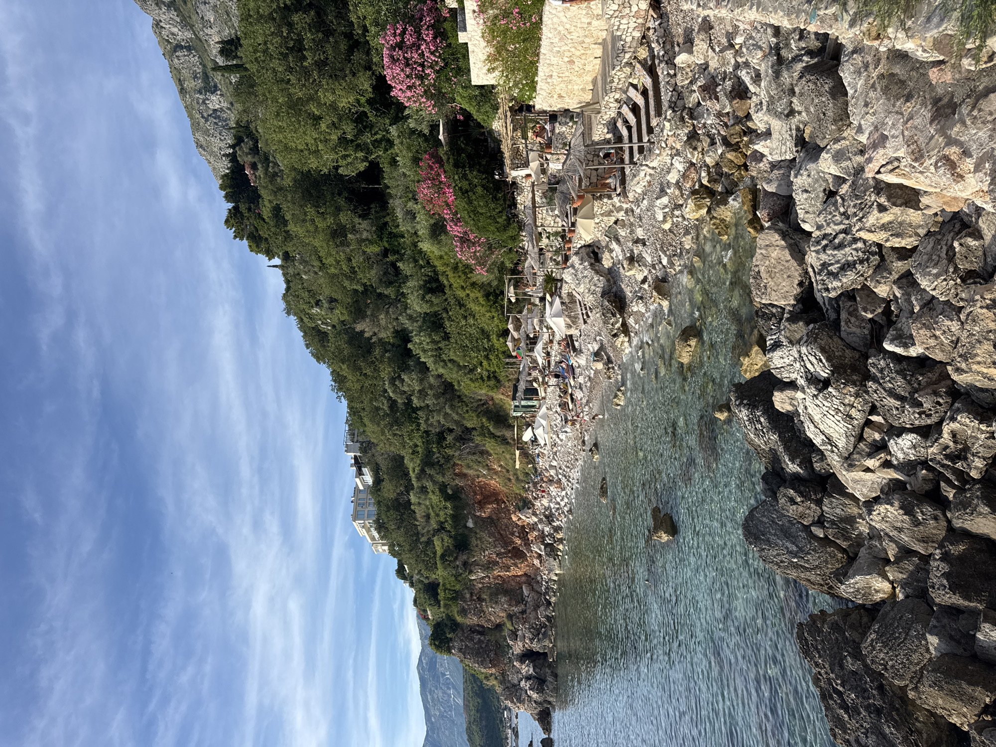 This image depicts a picturesque coastal scene in Rijeka Reževići, Montenegro. The foreground is dominated by large, weathered rocks of varying sizes and textures, bordering a crystal-clear, turquoise sea. The water is calm, with small waves gently lapping at the shore.
The middle ground features a rocky beach dotted with numerous sun loungers and straw umbrellas, indicating a recreational area. Several people are visible, relaxing on the loungers or walking along the beach. Behind the beach, a stone wall and terraces rise up the hillside, adorned with lush green vegetation and vibrant pink oleander flowers. Further up the incline, dense green trees cover the slope, leading to the base of a rugged, grey mountain.
In the upper portion of the image, a modern, multi-story white building with large windows is perched on the hillside amidst the greenery. The sky above is a clear blue with wispy white clouds, suggesting fair weather. The overall impression is one of a serene and beautiful seaside resort.