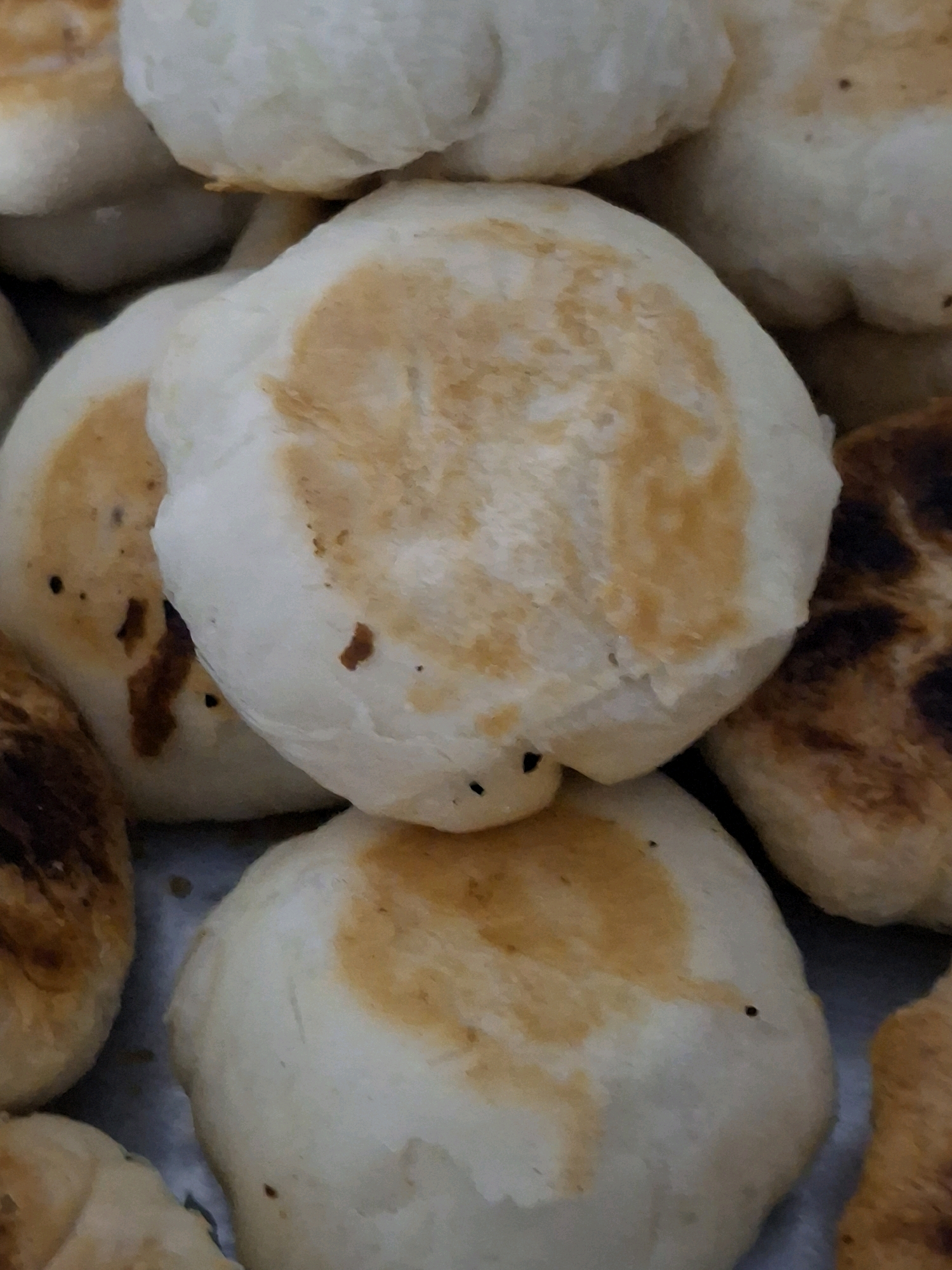 This is a close-up, overhead shot of several golden-brown and white baked goods, likely pastries or buns, arranged in a shallow container or on a flat surface. The lighting is soft and diffuse, highlighting the textures of the baked items. Some of the pastries have lighter, doughy centers with darker, toasted areas, suggesting they may have been pan-fried or baked in a way that created uneven browning. Small dark specks are visible on the surfaces of some of the pastries, which could be poppy seeds or other small inclusions. The background is a blur of similar baked goods and a metallic-toned surface that appears to be the container. There are no people or discernible environmental features of Lumajang, Indonesia, visible in the image.