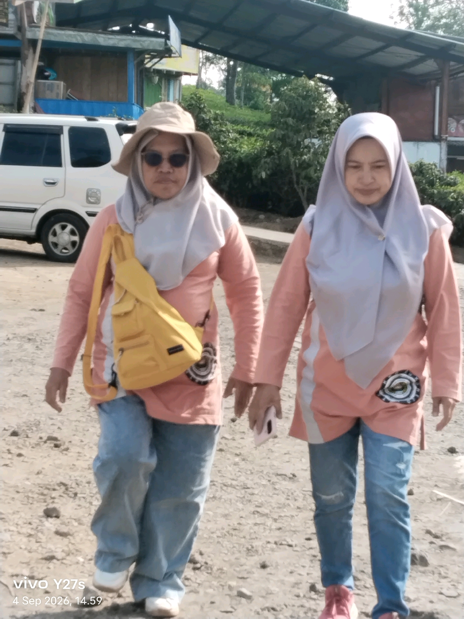 Two women are walking outdoors on a dirt path in what appears to be a rural or semi-rural area. The woman on the left is wearing a light-colored wide-brimmed hat, sunglasses, a light pink long-sleeved shirt, a light gray hijab, and light blue jeans. She has a yellow crossbody bag slung over her right shoulder. The woman on the right is wearing a light pink long-sleeved shirt with white paneling on the sides, a light gray hijab, and light blue jeans. Both women are facing forward and walking. In the background, there is a white SUV, some structures with a slanted roof, and vegetation, including trees. The overall scene suggests a casual outdoor activity.