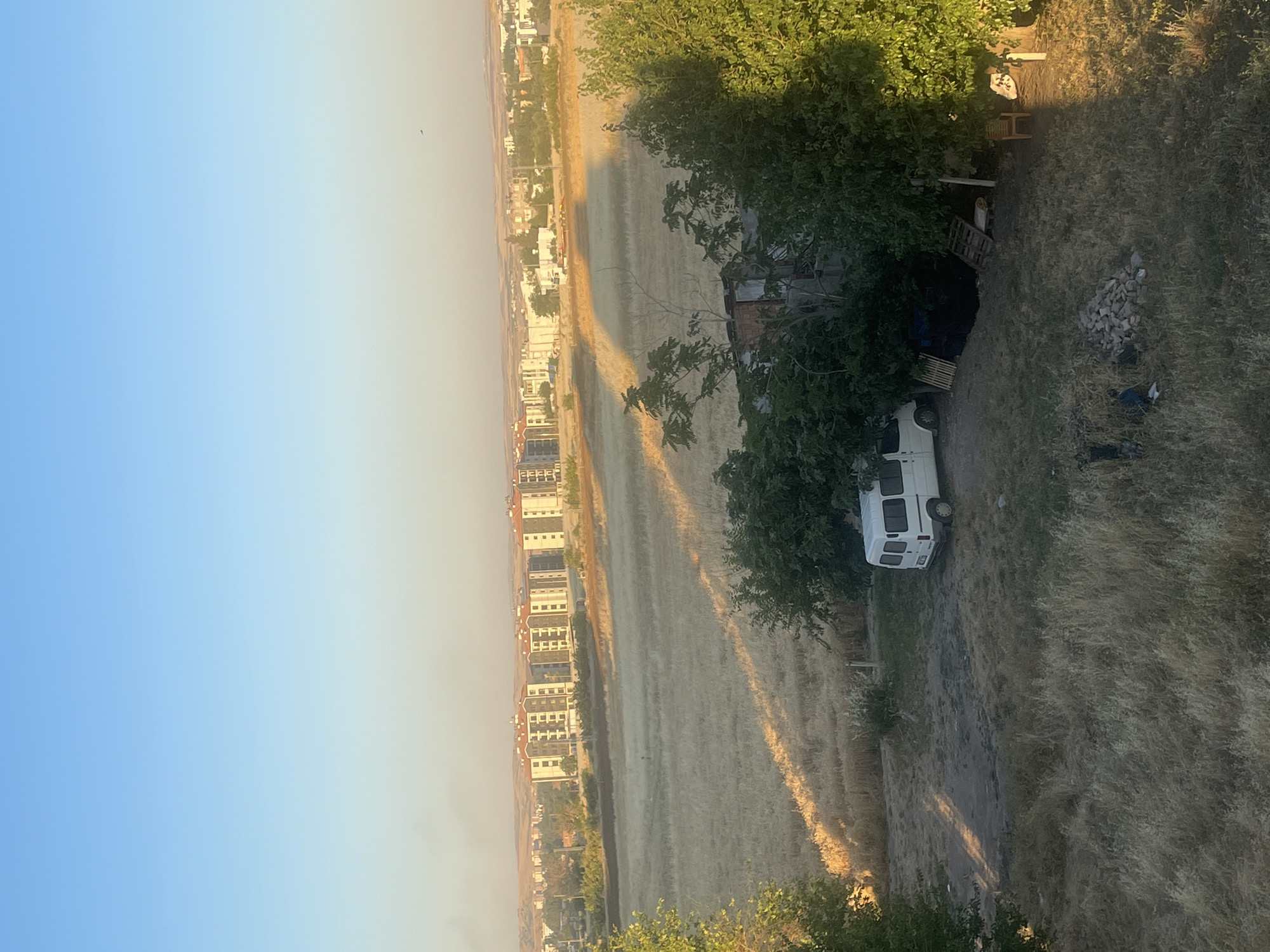 A high-angle, full shot captures a sunlit landscape with a city skyline in the background. The foreground features dry, tall grass and scattered debris, including a pile of grey stones, a dark tarp, and wooden pallets. A white van is parked in a dirt area surrounded by trees. In the mid-ground, a vast field of dry, golden stalks stretches towards the city. The background showcases a row of apartment buildings with red-tiled roofs, typical of Adıyaman, Turkey, under a clear, pale blue sky. The scene is illuminated by the warm, golden light of what appears to be early morning or late afternoon, casting long shadows across the field and ground.