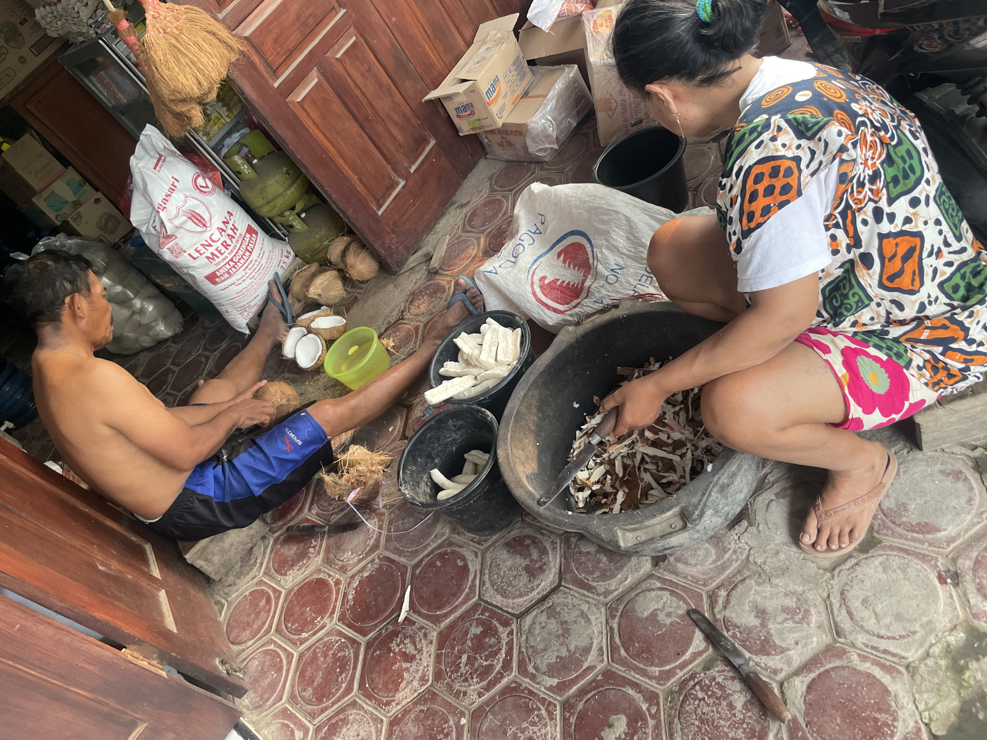 The image depicts an outdoor scene in what appears to be a residential area, possibly in Parakan, Indonesia, with a weathered wooden door and red hexagonal paving stones on the ground. Two individuals are engaged in preparing food, likely cassava.
In the foreground, a woman with dark hair is seated, wearing a white t-shirt with a colorful, patterned overshirt and a pink and green floral skirt. She is actively using a knife to shred what appears to be cassava into a large black bucket. Her feet are bare, and she wears a light brown flip-flop on her right foot.
Behind her, a man with dark hair, shirtless and wearing blue and black athletic shorts, is also working with coconuts. He is seated and holding a coconut in his left hand, with a knife near his right hand. Several halved coconuts and whole coconuts are scattered around him, along with a yellow plastic container. A bundle of coconut husks is visible near the man's feet.
Several large black buckets are present, one containing shredded cassava and another filled with large pieces of peeled cassava. A white sack labeled "PAGODA" and another sack with red lettering, possibly rice, are also visible. A green gas cylinder is situated next to the sacks. In the background, several cardboard boxes are stacked, and a broom made of natural fibers leans against the wooden door. The overall impression is one of a utilitarian preparation area, likely for food processing.