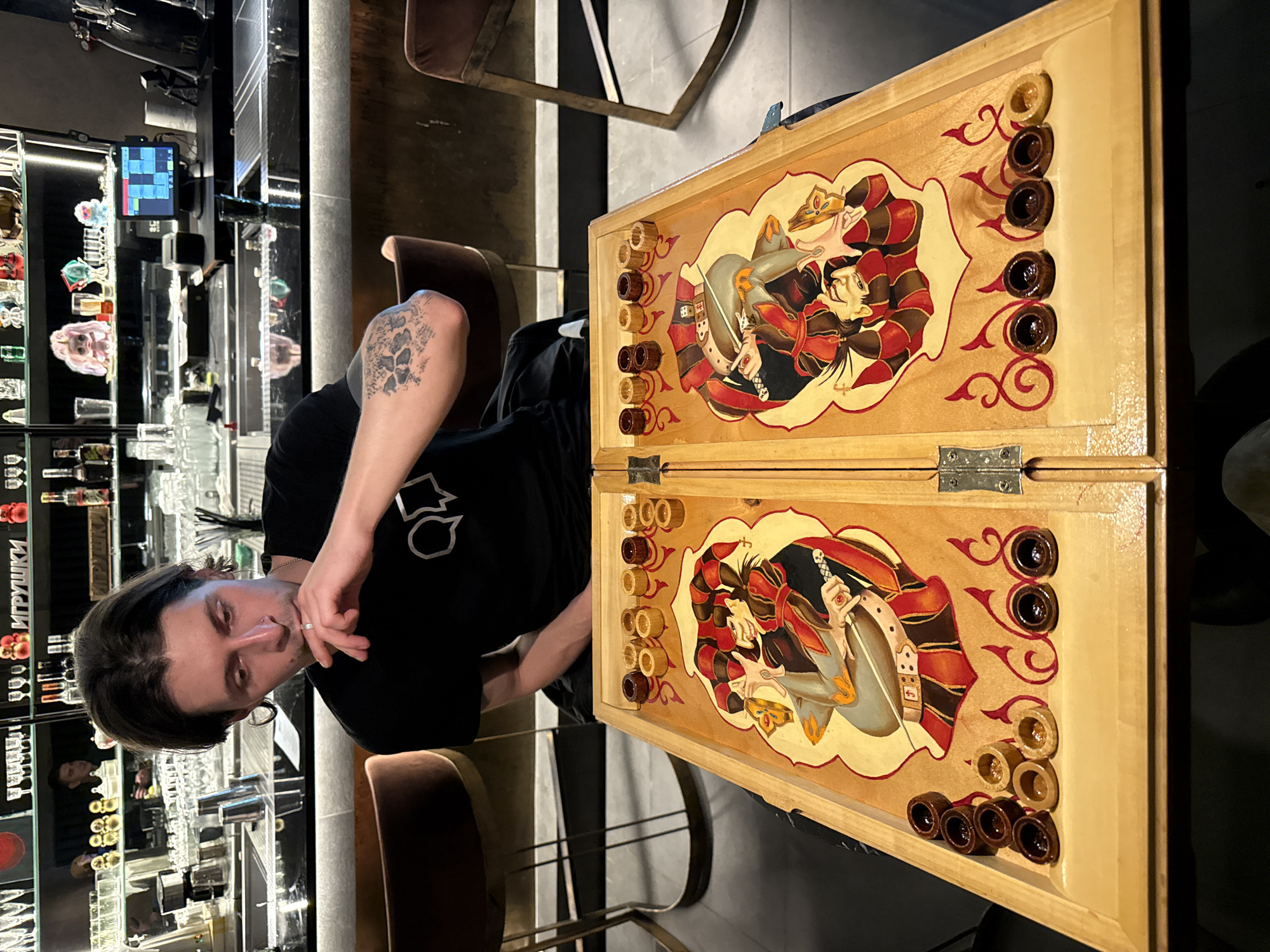 A man sits at a bar in Sochi, Russia, playing backgammon. He is wearing a black t-shirt and has a tattoo on his forearm. The backgammon board is decorated with a painting of two figures. The bar is well-lit and there are bottles of alcohol displayed behind the bar.