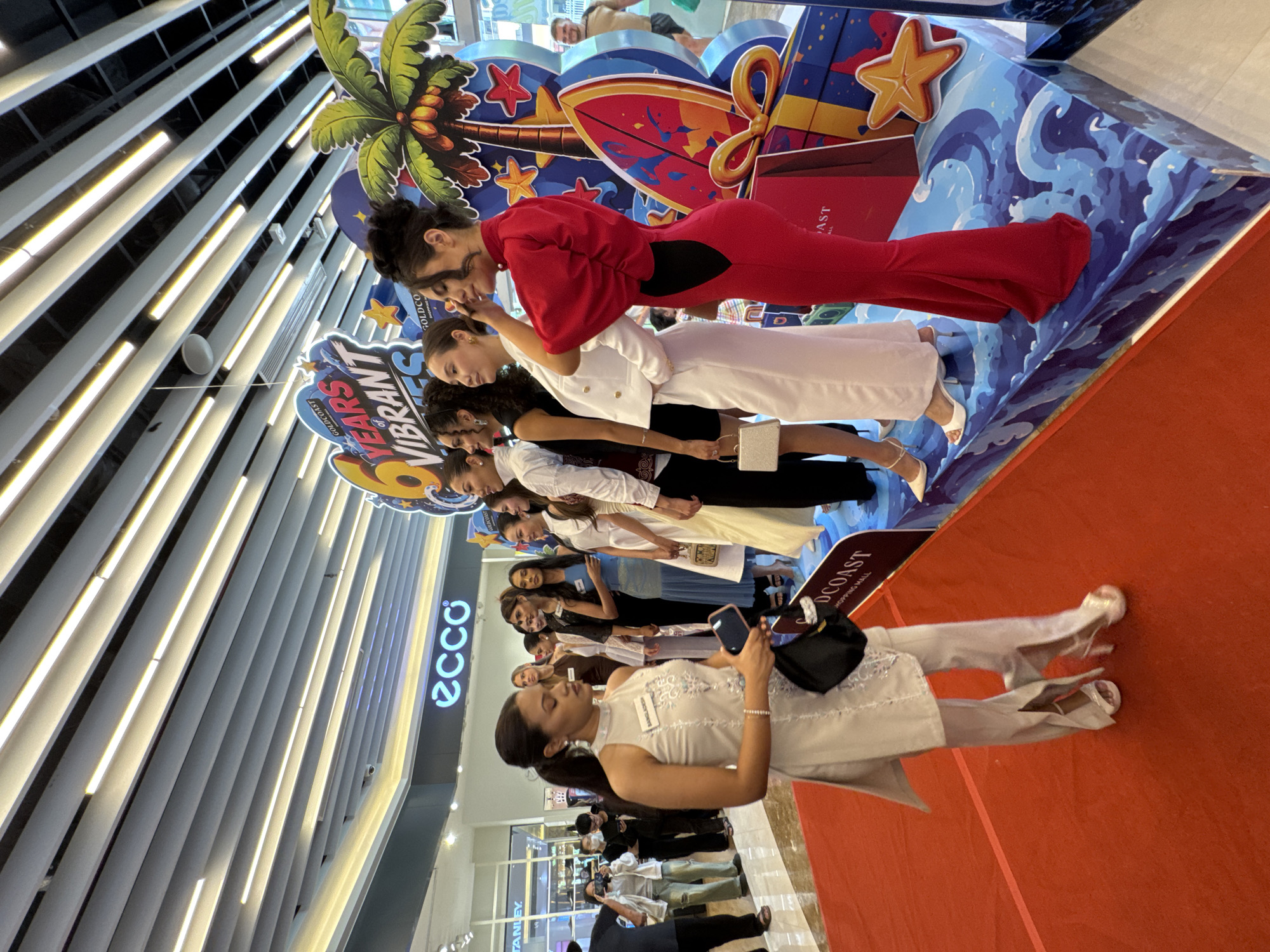 In Nha Trang, Vietnam, a group of women in formal attire are posing on a red carpet in what appears to be a shopping mall. A banner behind them reads "GOLDCOAST 6 YEARS OF VIBRANT". The women are of various ethnicities and are dressed in a range of colorful and elegant outfits, including a striking red gown and a white suit. One woman, wearing a light-colored ensemble, is in the foreground and appears to be addressing someone off-camera while holding a mobile phone and a black clutch. Another woman, in a red gown, is adjusting her hair, while a third woman, in a white suit, is also looking away. The background features a vibrant display with palm trees, surfboards, and gift boxes, suggesting a celebratory or promotional event. The "ecco" logo is visible on a sign in the background, indicating a potential partnership or sponsorship. The overall atmosphere is festive and glamorous.