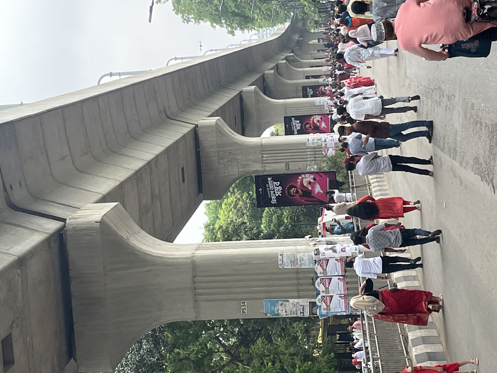 A long line of people walks along a street in Dhaka, Bangladesh, under an elevated concrete structure with pillars marked "P534". The crowd consists of diverse individuals, some wearing white shirts and caps, others in casual attire, and a few in red dresses. Numerous posters and advertisements are affixed to the pillars supporting the overhead structure. The street is lined with trees and foliage, and the sky is overcast. A person on a bicycle is visible at the edge of the frame.