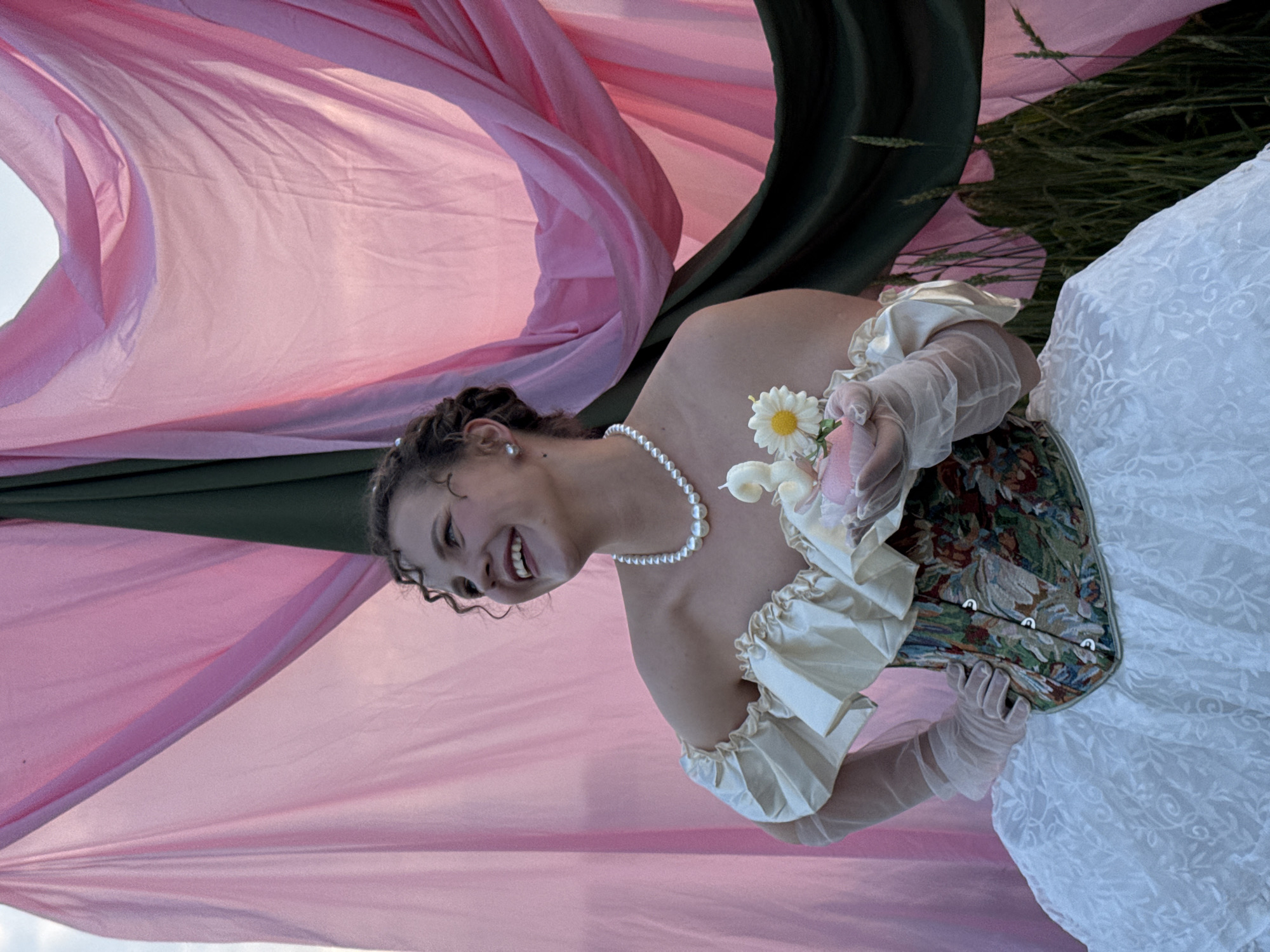 A young woman is pictured from the chest up, holding a small cake with a lit candle shaped like the number '2' and a daisy. She wears a white pearl necklace, a cream-colored off-the-shoulder top with puffy sleeves, a floral-patterned corset, and a white lace skirt. She also wears sheer white gloves. The woman is smiling and looking to her left, with curly brown hair. Behind her, a backdrop is made of draped pink and dark green fabric. The scene appears to be outdoors, with tall grass visible in the lower right corner. The location is in the city of Fok, Russia.