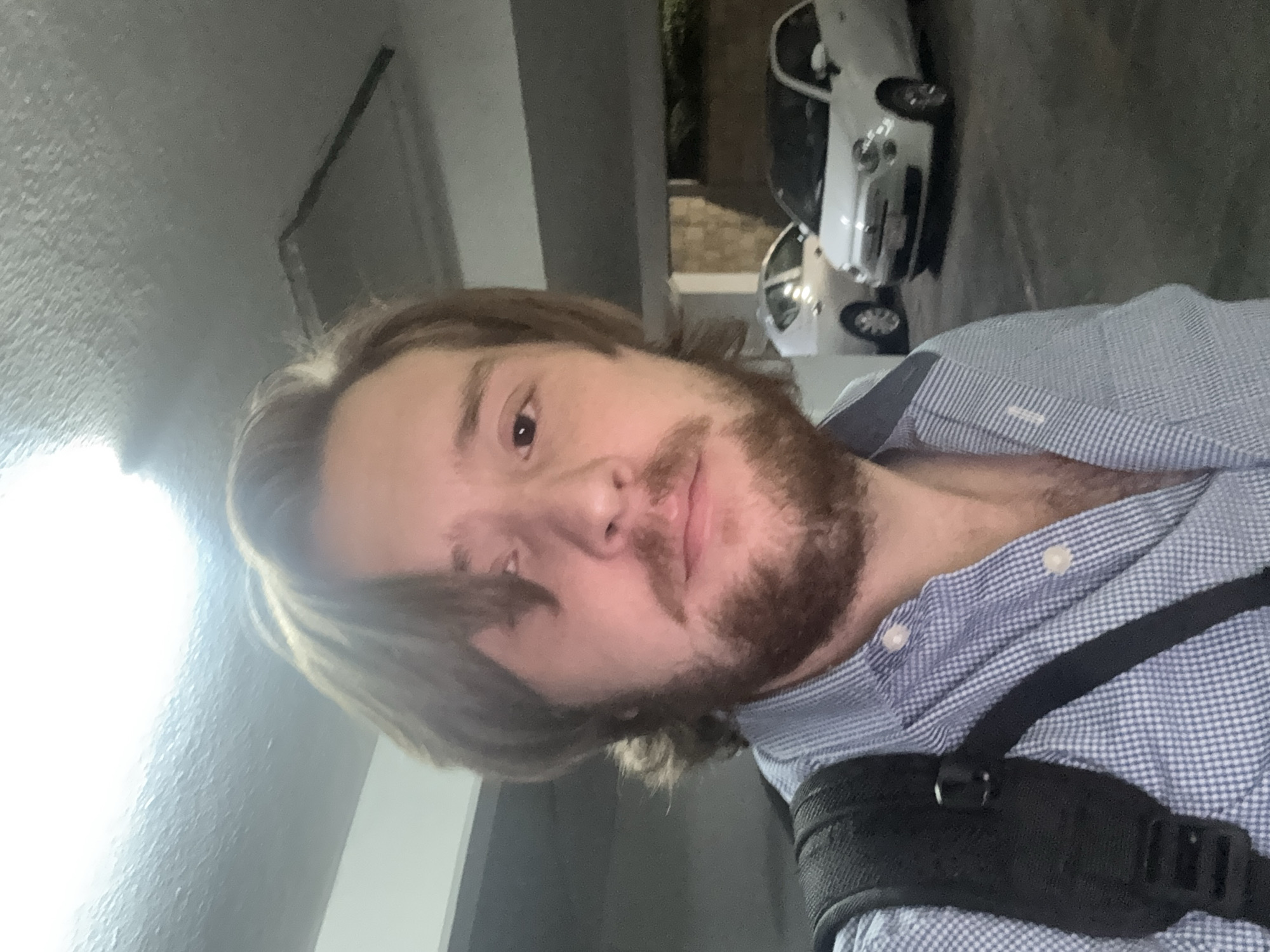A selfie of a person with brown hair and a beard, wearing a blue checkered shirt and a black strap. The background shows a garage with two white cars. Based on the visible environment and metadata if available, the location is presumed to be Mexico City, Mexico.