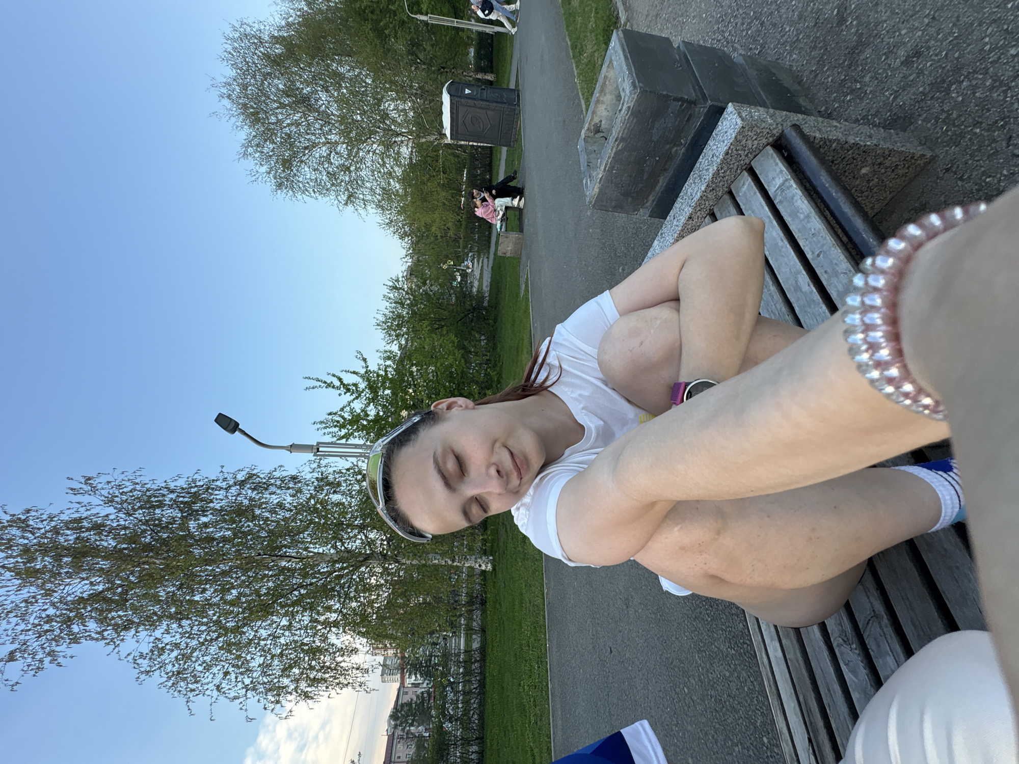 The image depicts a woman sitting on a wooden bench in a park-like setting. She is wearing a white t-shirt and has her legs drawn up to her chest. She has dark hair pulled back and is wearing sunglasses on top of her head. The woman's eyes are closed, suggesting she is resting. A bracelet is visible on her wrist closest to the camera.
The background shows trees, a walkway, and a portable toilet unit, indicative of a public park. Other people can be seen walking in the background. A building is also visible in the background.
Based on the background details, this photograph was most likely taken in the city of Perm, Russia.