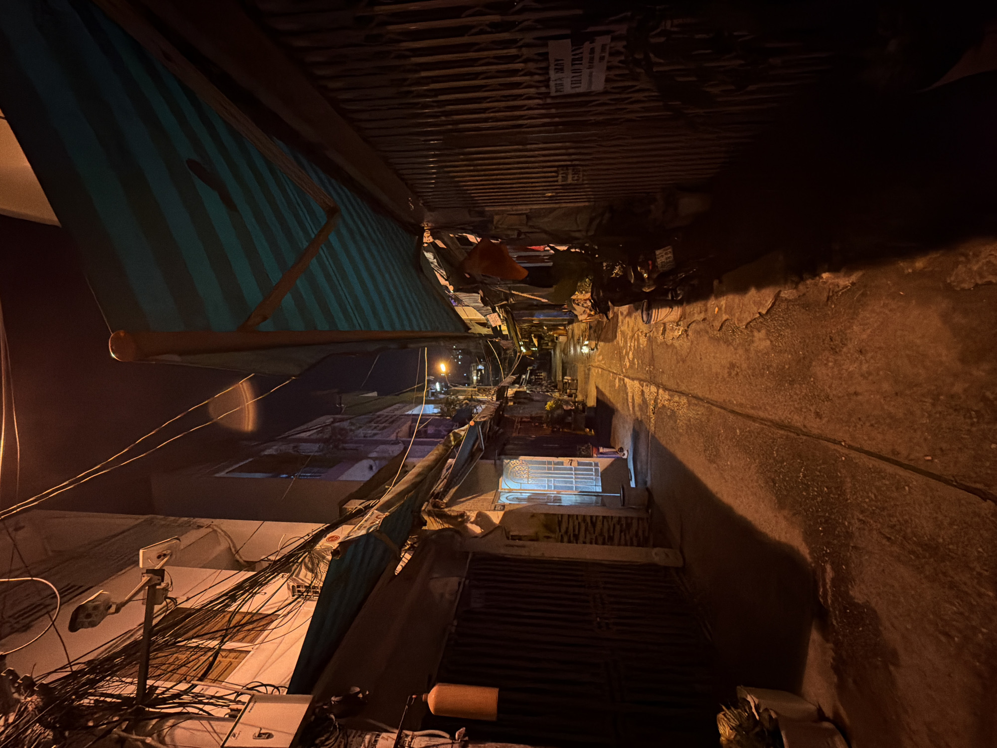 The image captures a narrow, bustling alleyway at night in Thủ Đức, Vietnam. The scene is illuminated by artificial lights, casting warm, yellow-orange tones and creating deep shadows.
On the left side of the alley, a prominent blue and white striped awning extends over a building, partially obscuring the lower levels. Above it, a dense network of tangled power lines crisscrosses, connecting to buildings and utilities. A streetlamp with two fixtures is visible amidst the wires, suggesting a source of illumination for the area.
Buildings line both sides of the alley, appearing to be multi-story structures. Many feature closed metal roller gates or grilles, indicating closed shops or residential entrances. The walls show signs of wear and age. Further down the alley, distant lights hint at the continuation of the street or other buildings.
The right side of the alley features a grimy, textured wall, possibly concrete or plaster, with distinct shadows stretching along its surface. What appears to be a small motorbike or scooter is parked near the right wall, partially obscured by the darkness, with some items like a small bucket or canister nearby. An orange object, possibly a gas cylinder, is seen closer to the foreground on the left side, under another awning or structure.
Visible text on a white sign attached to a corrugated metal ceiling or awning reads "PHÚC KHƠI" and "VĂN TRANG," likely names of businesses or individuals. The overall impression is that of a dense, urban environment characterized by its tight spaces, overhead wiring, and nighttime activity.