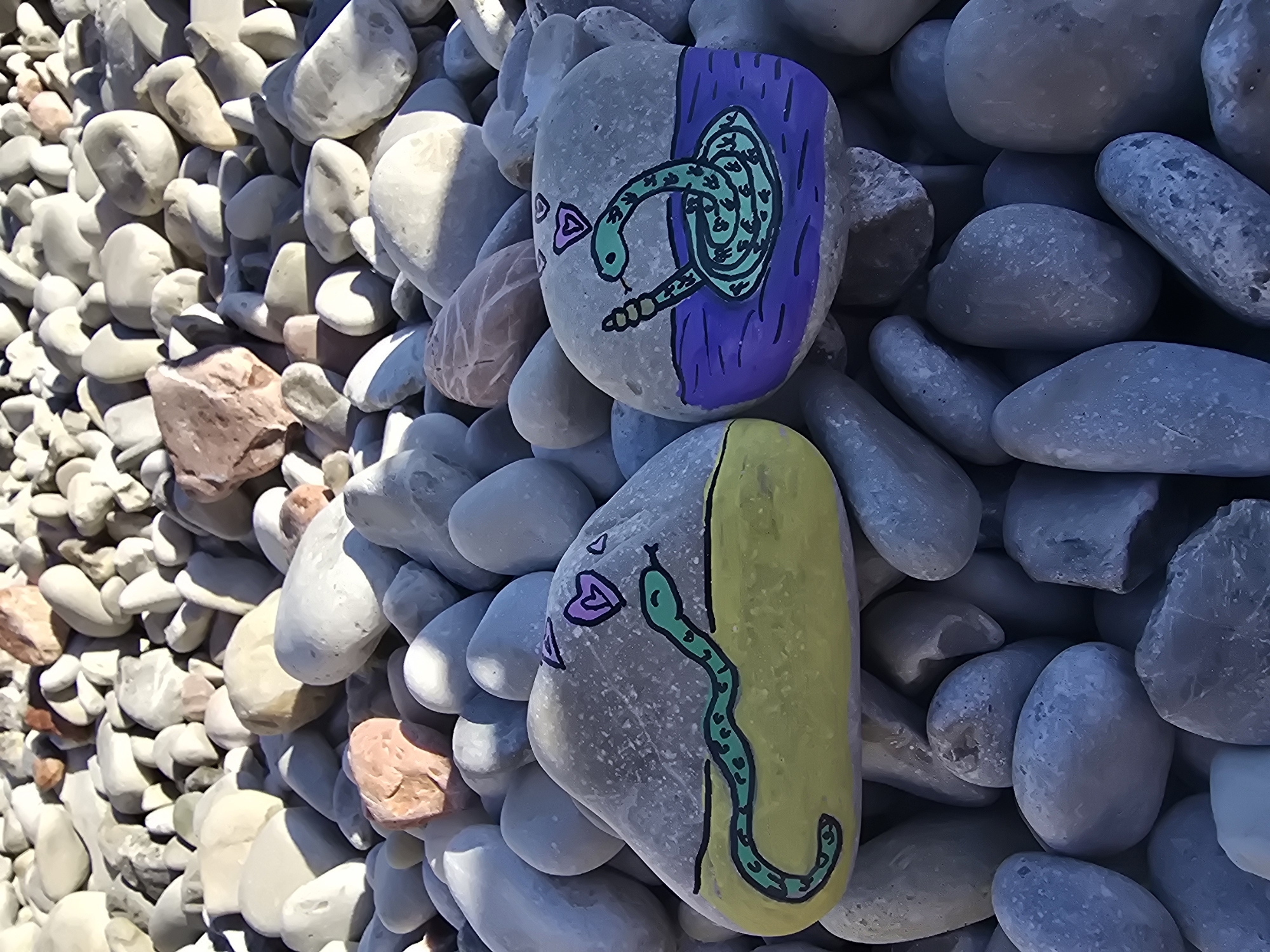 The image displays two rocks painted with cartoonish snakes, situated on a bed of grey and pinkish-white pebbles. The rock on the left features a green snake with black markings, lying on a yellow background, with two purple hearts above it. The rock on the right shows a similar green snake with black markings, coiled in purple water, also with two purple hearts above it. The pebbles suggest an outdoor environment, possibly a beach or riverbed. The location is identified as Mirište, Montenegro.