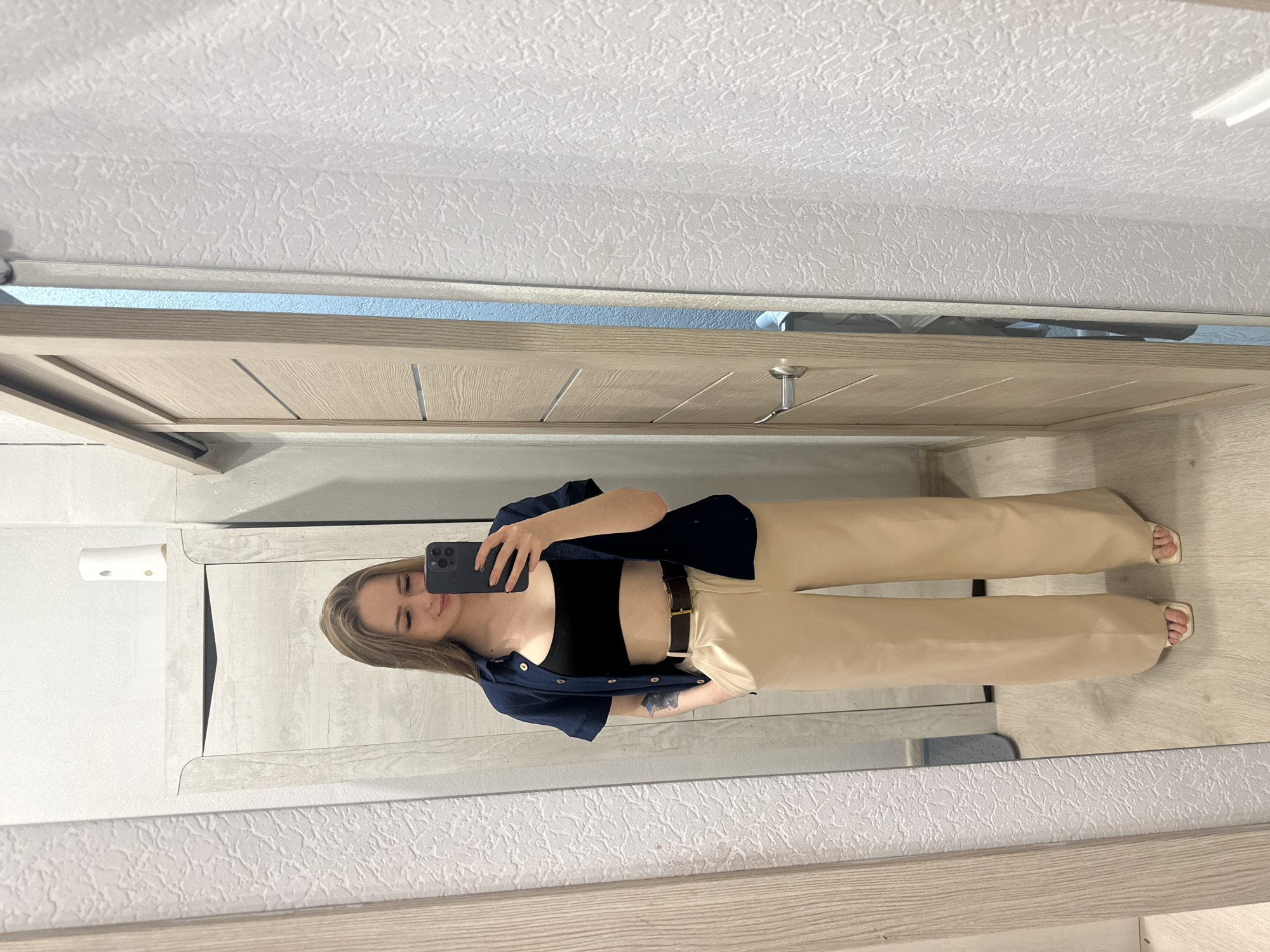 A woman stands in front of a mirror, taking a photo of herself with a smartphone. She wears a black top, tan pants with a black belt, and a dark blue button-down shirt. The background includes a door, a wall, and a ceiling, all painted in neutral tones. The location appears to be inside a residential building.
