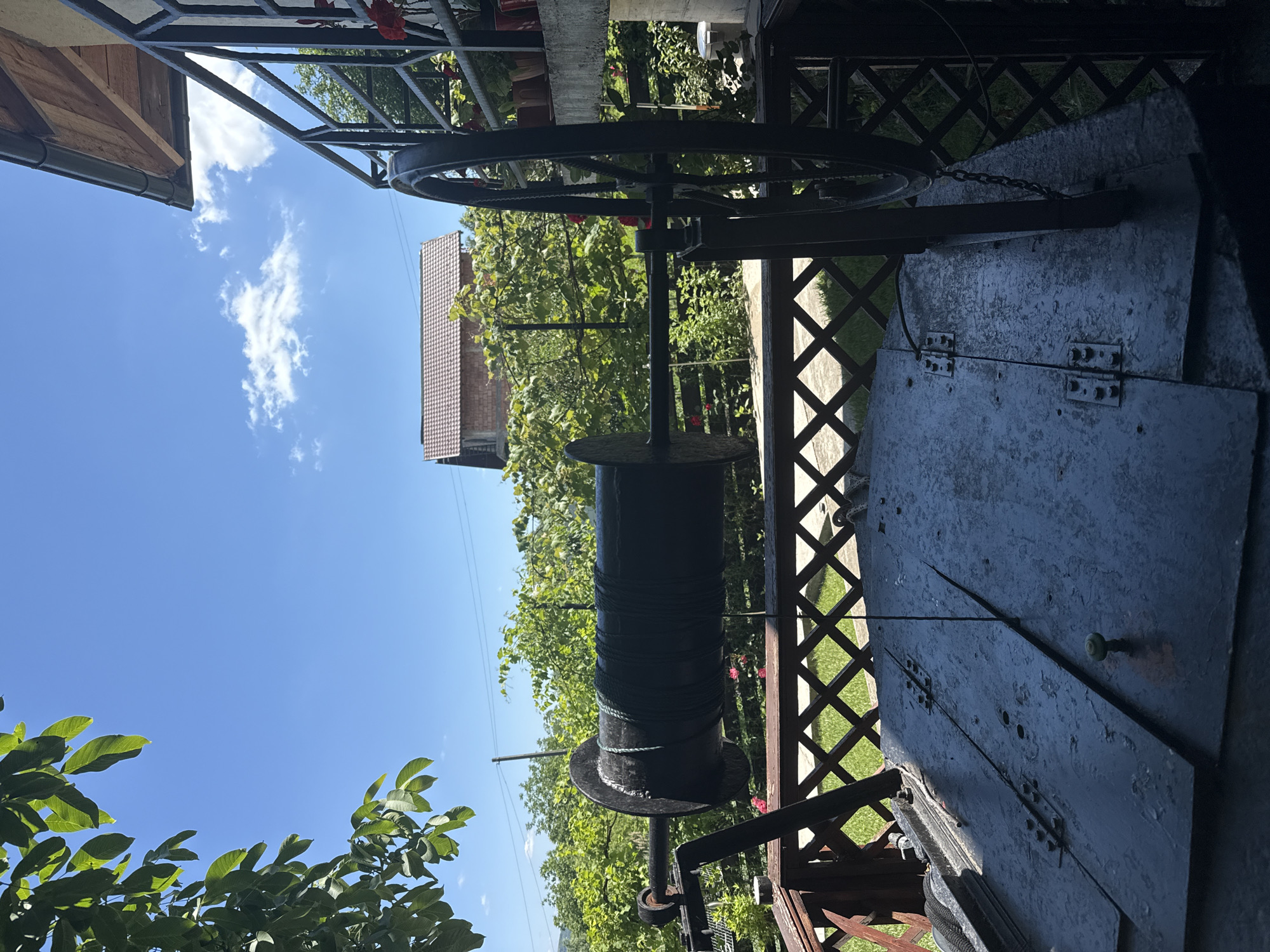 The image shows an old-fashioned well with a large wooden reel, a crank mechanism, and a metal cover. The well is located in a rural setting, possibly a backyard or garden, with lush greenery and a clear blue sky. In the background, there is a house with a tiled roof and a fence. The overall atmosphere is rustic and peaceful.
