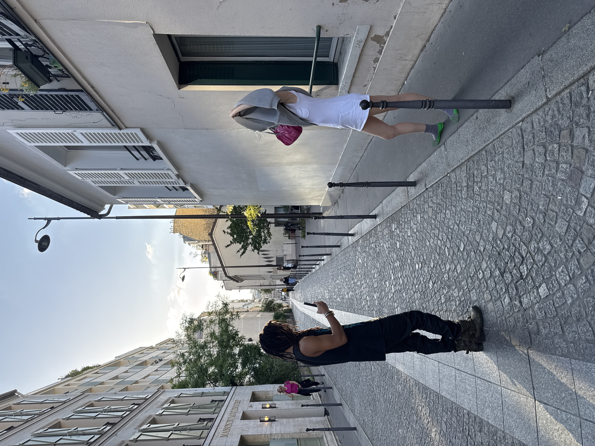 The image captures a street scene in Paris, France. The perspective is unusual, as the image is rotated ninety degrees. Cobblestone paving covers the street, flanked by buildings on either side.
On the right side of the image, a woman stands on the sidewalk. She wears a white dress, a gray jacket, and green sneakers. She carries a pink purse. She appears to be leaning against a metal post.
On the left side of the image, a person with dreadlocks is lying on the cobblestone street, looking at a cell phone. They are dressed in dark clothing. Further down the street, another person is standing on the sidewalk near a metal post.
Buildings line both sides of the street, with architectural details like shuttered windows visible. A street lamp is visible on the left. The sky is light blue. The street appears to be narrow, leading into the distance. A sign that reads "MAISON DU LEVERT" is visible on the facade of the building to the left.