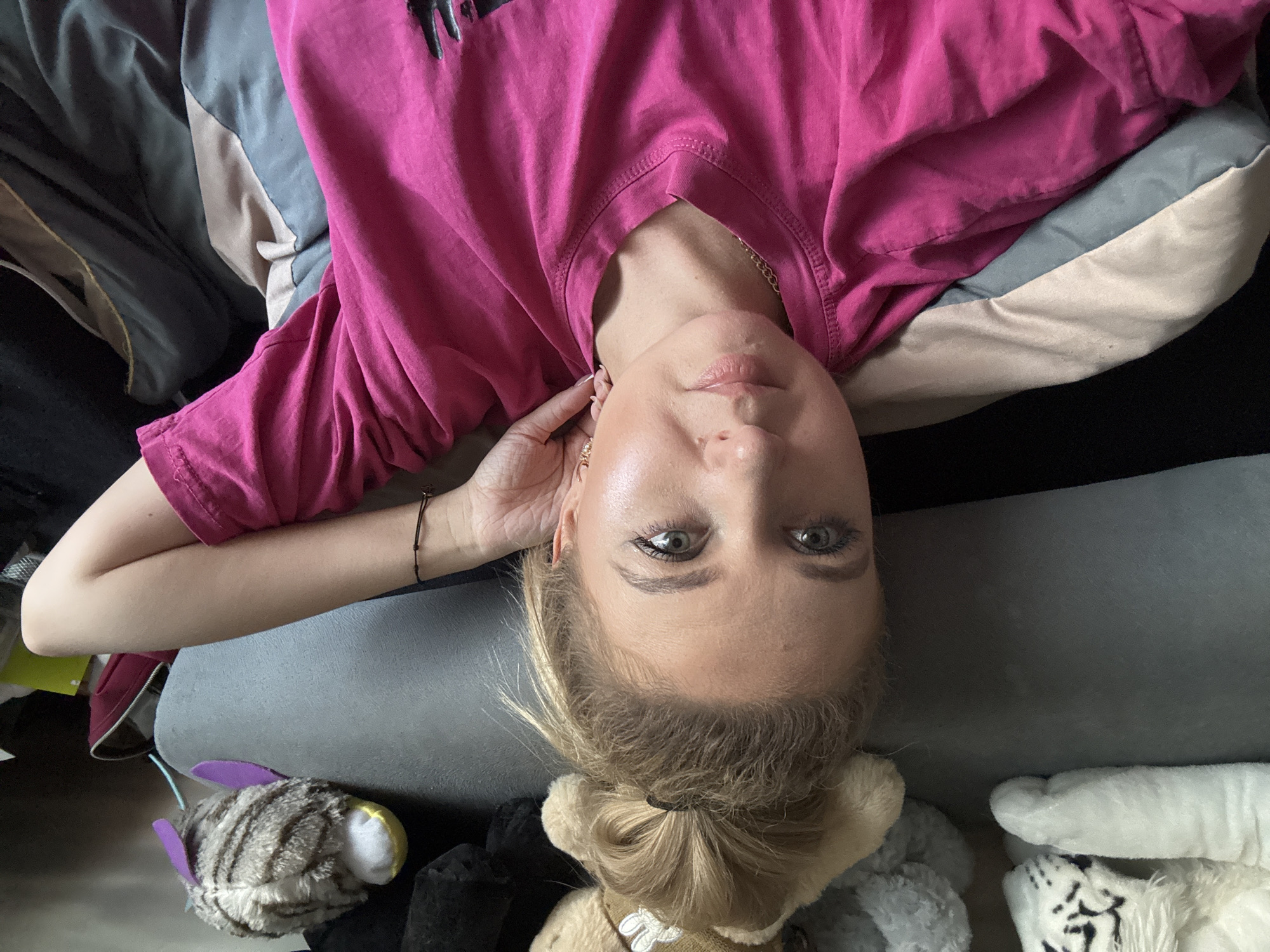 A woman is lying on a bed, with her head tilted to the side, as if taking a selfie. She is wearing a pink shirt and has her hair pulled back. There are several stuffed animals around her, including a striped cat, a teddy bear, and a white tiger. The room is dimly lit, suggesting it might be nighttime or a cloudy day. The woman's expression is relaxed and a little playful. There is no visible text in the image.