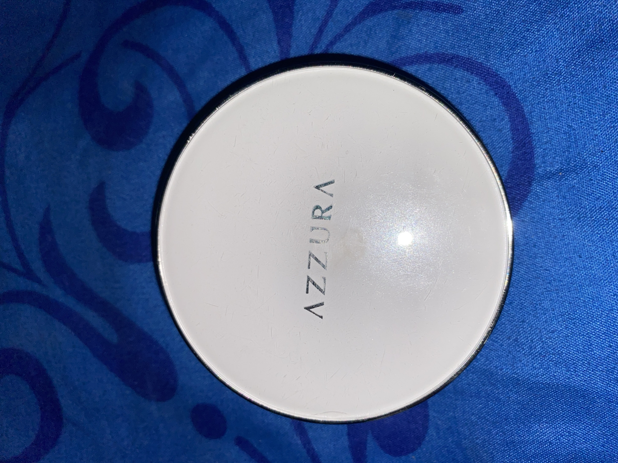 A round, white compact sits on a blue, patterned fabric surface. The compact has the word "AZZURA" embossed in silver lettering on its lid, with visible scratches on the surface. The background fabric has a swirling, abstract pattern in darker blue. The location is not discernible from the image.