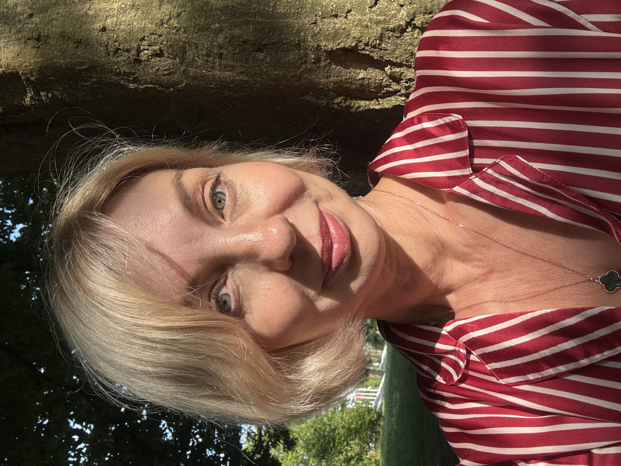A woman with blonde hair and light blue eyes takes a selfie outdoors. She is wearing a red and white striped shirt and a gold necklace with a dark clover pendant. The woman is standing next to a tree trunk. In the background, there are green trees and a hint of a white structure. The image appears to be taken in a park or garden. There is no specific indication that this location is in the city of Sochi, Russia.