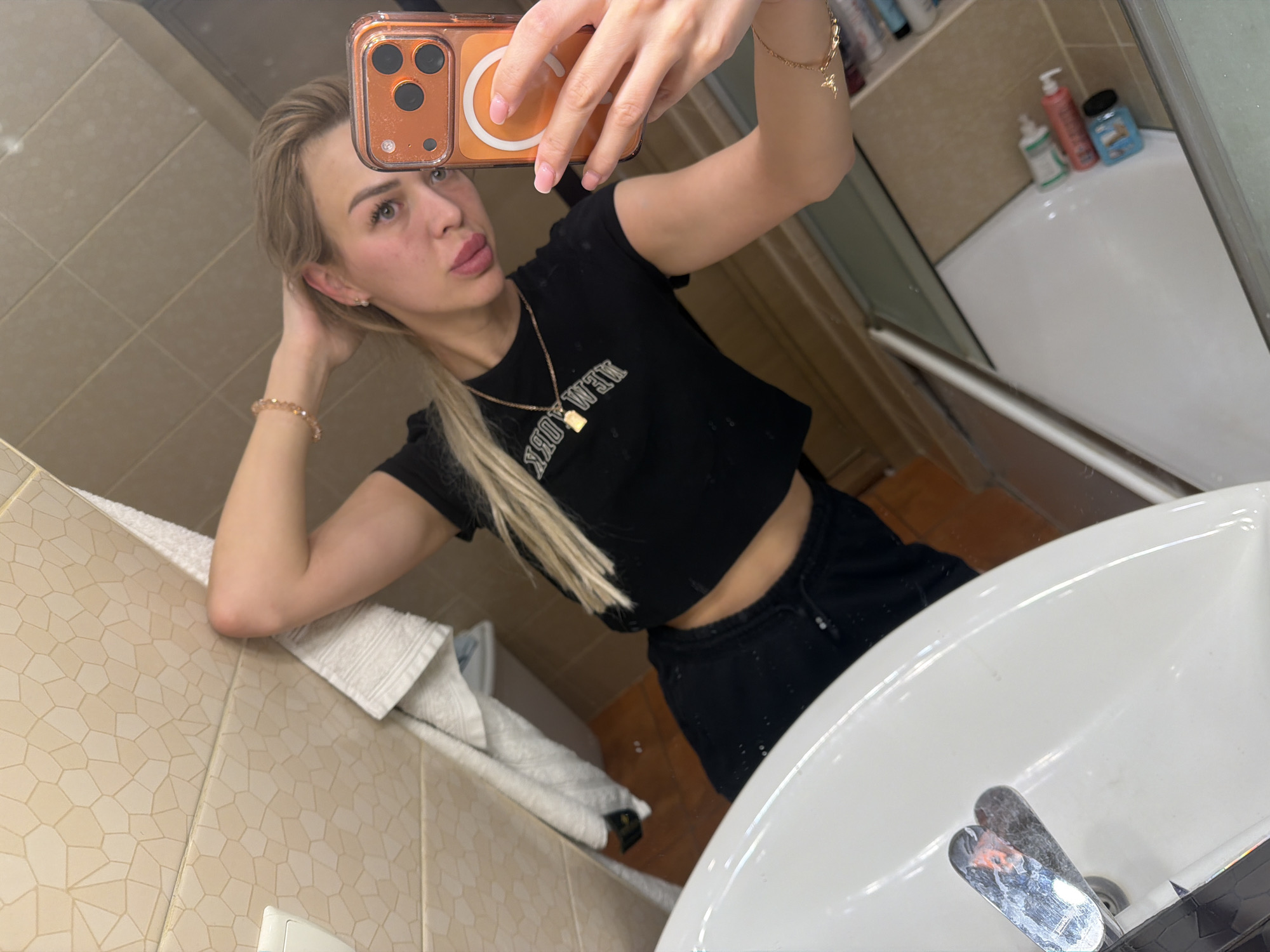 A young woman with blonde hair, wearing a black cropped t-shirt and black pants, is taking a mirror selfie in a bathroom. She is holding her phone, which has an orange case, in her right hand, and her left hand is behind her head. She is wearing jewelry on her wrist and neck. The bathroom has tiled walls and a white sink. The overall environment appears to be a typical residential bathroom.