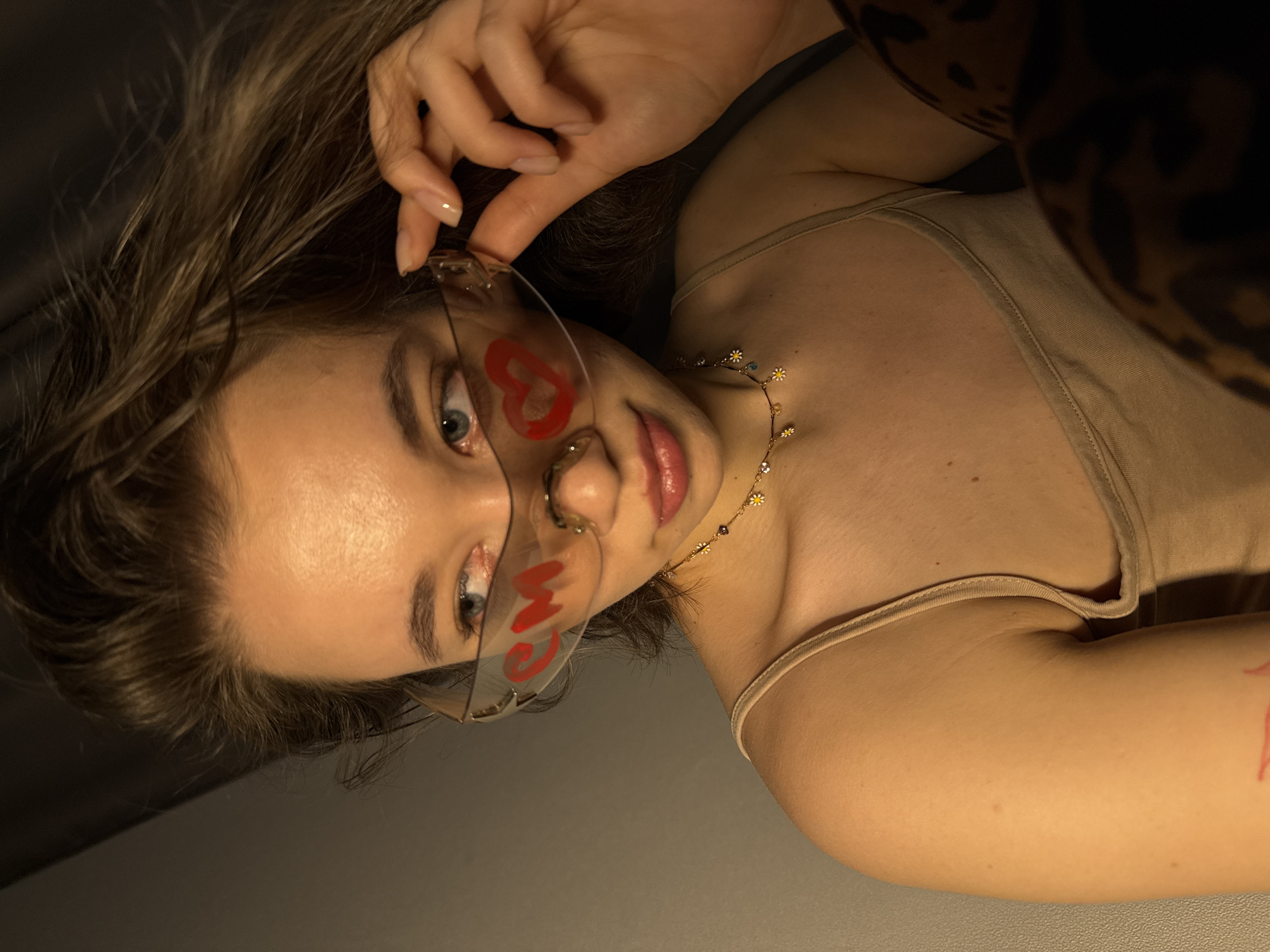 The image presents a close-up, angled portrait of a young woman. She is positioned as if lying down, with her head tilted, and her gaze directed towards the camera. She has fair skin, blue eyes, and light brown hair that cascades around her head.
She is wearing a light beige or nude-colored tank top, and a delicate gold-toned necklace with small daisy-like charms and clear beads adorns her neck. Her right hand is gently raised, with fingers near her hair and the frame of her clear, rimless glasses.
A distinct feature of the image is the red drawing on her glasses. A red heart shape is visible over her left eye (viewer's right), and the letters "SW" are drawn in red over her right eye (viewer's left). Her lips are slightly parted, hinting at a subtle smile.
The setting appears to be indoors, characterized by soft, warm, and somewhat dim lighting that creates a focused, intimate atmosphere. The background is largely dark and indistinct, ensuring the woman remains the central focus. A dark, possibly patterned (e.g., leopard print) fabric is partially visible on the right side of the frame, adding a touch of visual interest.
The woman's expression is calm and direct, conveying a thoughtful or engaging mood. No specific location cues within the image itself indicate the context of Noviy, Russia; the scene is consistent with a private indoor space. The lighting suggests it could be evening or night, or a controlled lighting environment.