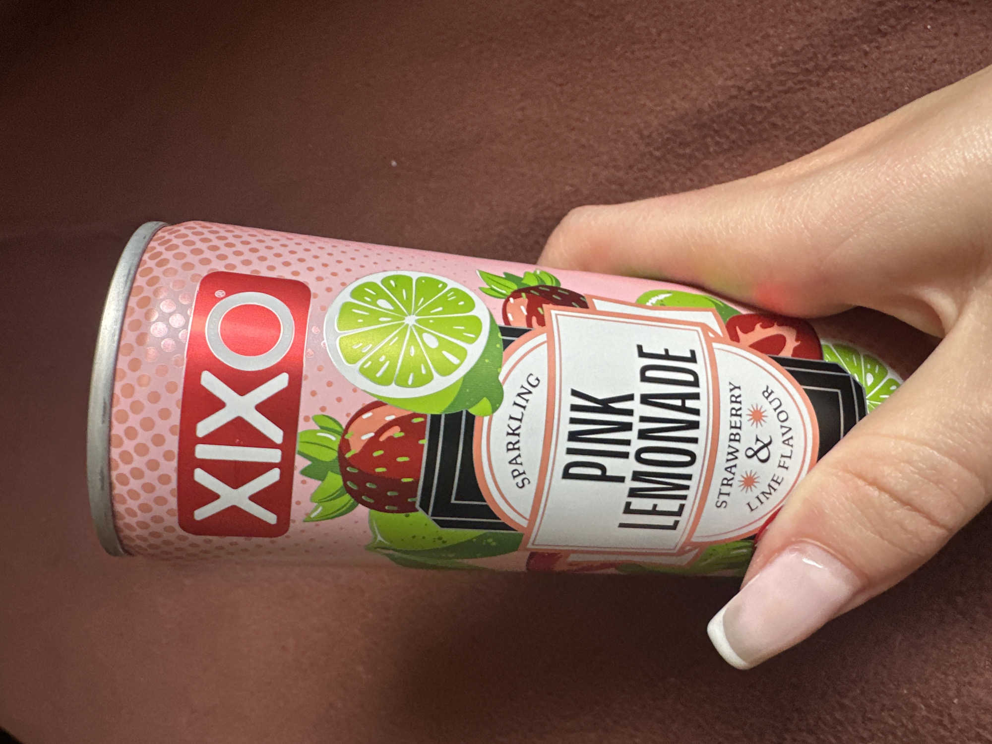 A close-up shot features a hand holding a pink Xixo Sparkling Pink Lemonade can, positioned against a blurred brown background. The can's design includes images of strawberries and limes, along with the text "Pink Lemonade," "Sparkling," and "Strawberry & Lime Flavour." The hand holding the can has manicured nails with a French tip design. The image does not provide any context as to the location being Sofia, Bulgaria.
