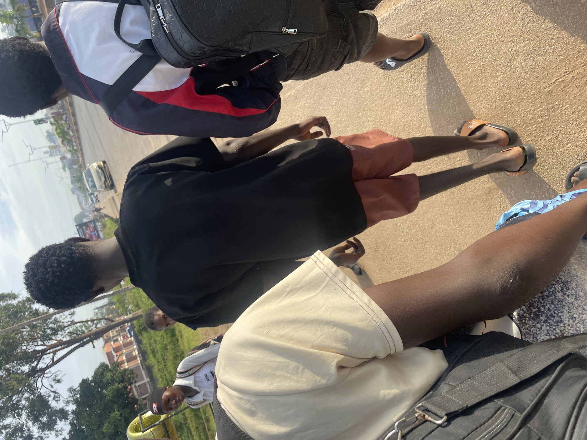 The image depicts a group of young people walking on a paved surface, likely a sidewalk, next to a road in Barakin Lerawi, Nigeria. The foreground shows the back of a person wearing a light-colored t-shirt and a black backpack. To their right, another person walks away from the camera, wearing a black t-shirt and orange shorts, with a black backpack visible. Further to the right, a third person is seen from the back, wearing a jacket with red, white, and black sections, and a black backpack. In the background, there are trees, buildings, a yellow circular object, and vehicles on the road. The scene appears to be outdoors during daylight, with the sun casting shadows. The overall impression is of individuals moving along a street in a developing urban or semi-urban area.