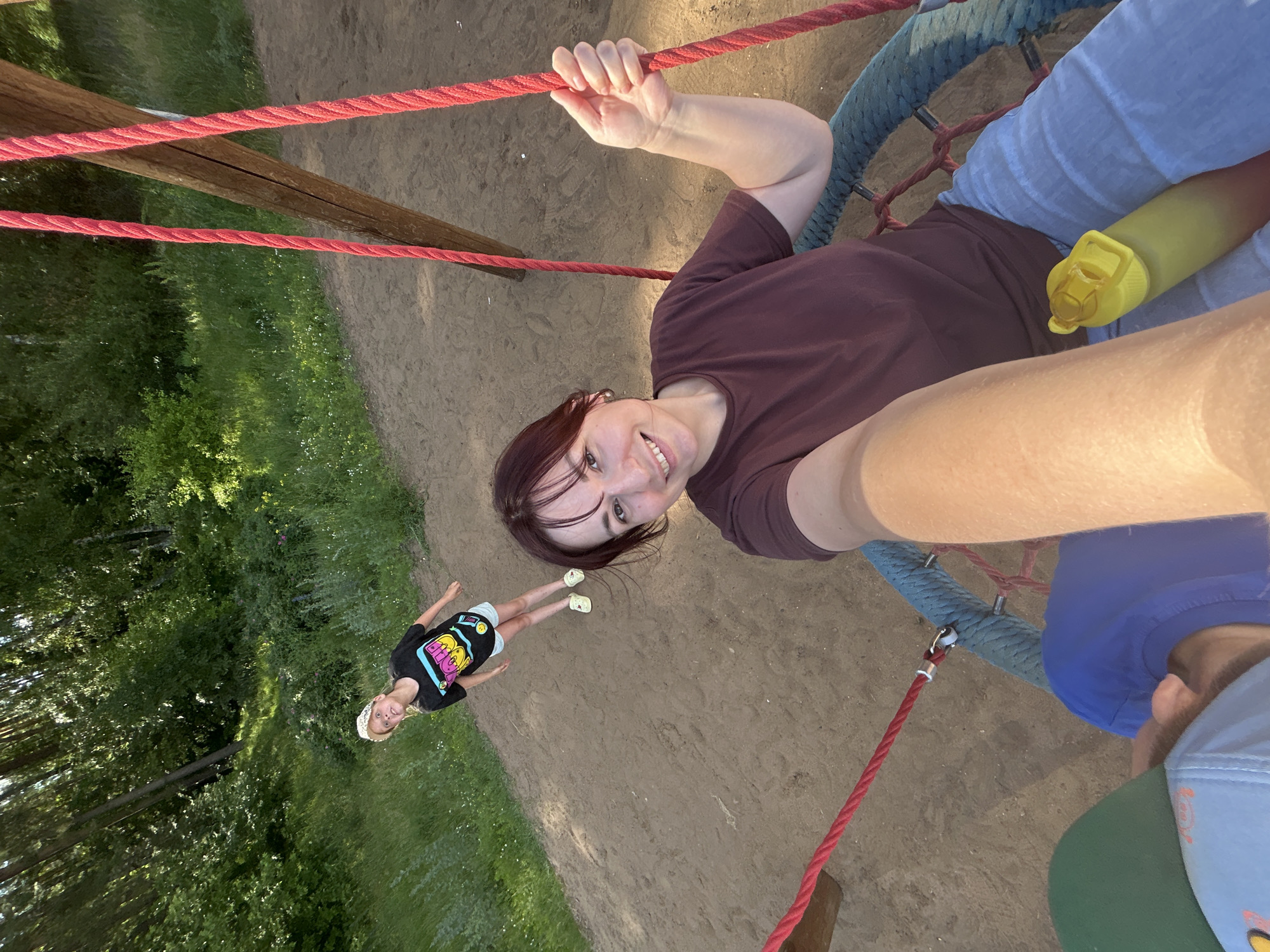 The image shows two individuals, an adult female and a child, on a playground in Chaikovsky, Russia. The adult female is in the foreground, smiling and holding onto the rope of a swing. She is wearing a brown t-shirt and blue jeans. A yellow water bottle is visible on her lap. The child, a young girl, is in the background, standing and looking up. She is wearing a black t-shirt with colorful graphics and white shorts. The playground appears to be made of sand, with trees and grass in the background. The sun is shining, casting shadows on the ground.