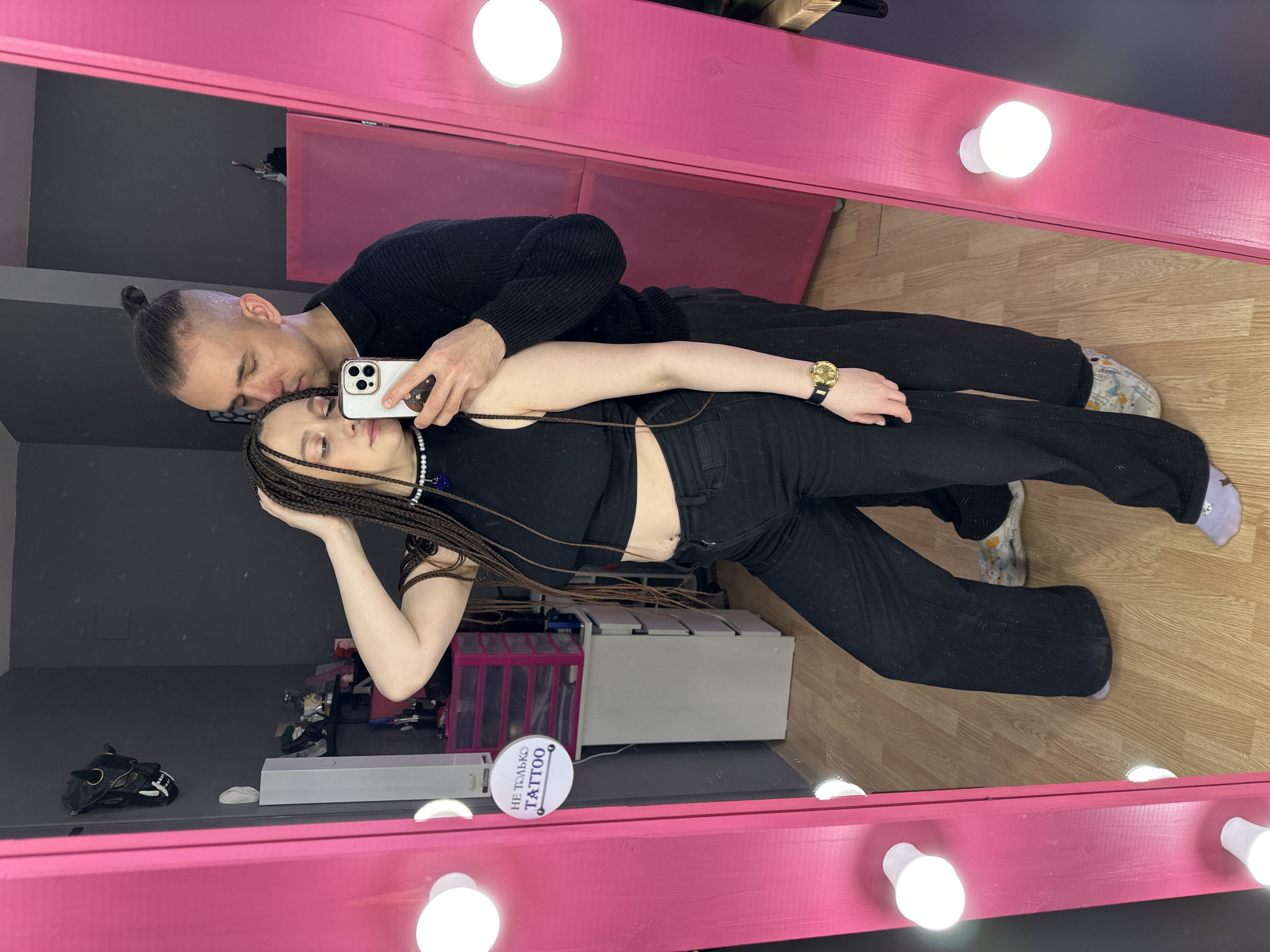 An indoor mirror selfie captures two individuals, a male and a female, in what appears to be a studio or salon in Saratov, Russia, indicated by a circular sign reading "НЕ ТОЛЬКО TATTOO" (Not Only Tattoo).
The female subject, standing in the foreground, has long dark brown micro braids and is wearing a black sleeveless crop top, black wide-leg pants, and light purple socks. A white beaded choker with a blue pendant adorns her neck, and a gold-toned watch with a black band is on her left wrist. She has a belly button piercing visible. Her right hand is raised to her braided hair near her temple, and her eyes are partially closed.
The male subject stands behind the female, wearing a dark long-sleeved ribbed sweater and dark pants with light-colored, patterned slip-on shoes. His hair is styled with shaved sides and a top knot. His face is positioned against the female's temple, with his eyes closed. He holds a white smartphone, which is used to capture the mirror selfie, obscuring part of his face and the female's left cheek.
The background, reflected in the mirror, features dark gray walls and a light wood laminate floor. A large vanity mirror with a thick pink wooden frame and multiple spherical white light bulbs dominates the foreground. To the left of the reflection, a black horned mask is mounted on the wall above shelving units containing various items and bottles. To the right of the reflection, a white cabinet with drawers is topped by a pink multi-drawer storage unit. The "НЕ ТОЛЬКО TATTOO" sign is white with black text and mounted on the wall to the left of the pink storage unit.