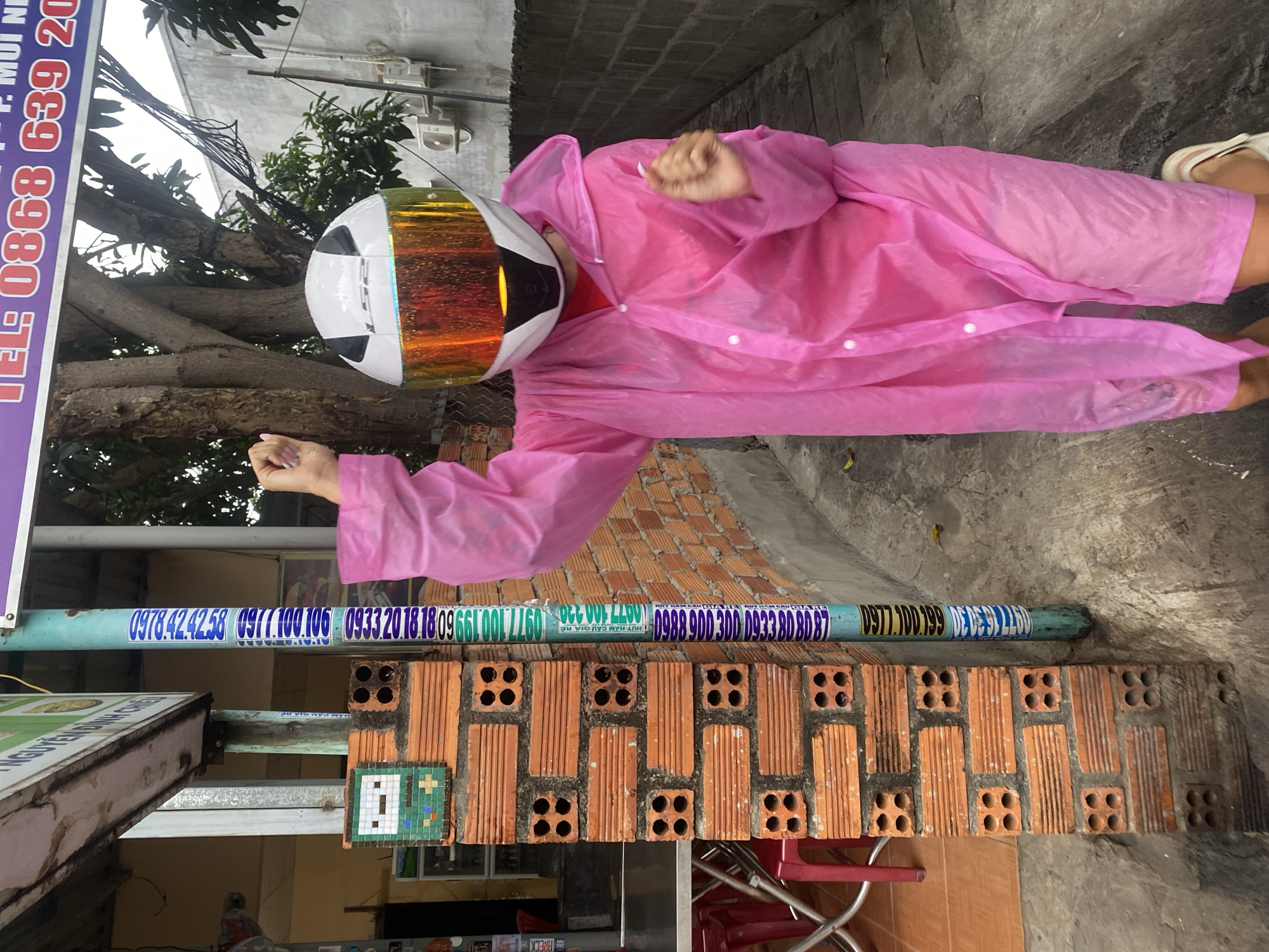 A person in a pink raincoat and helmet stands outside. The person is wearing white sandals. Behind them, a low brick wall is partially visible in front of a grey concrete wall. A pipe painted with various phone numbers is attached above the brick wall. Part of a sign is visible on the left with the text "EL: 0868 639 20" and "NGUYEN DINH CHIE". The location is Phu Thuy Ward, Vietnam, based on the phone numbers in the image.
