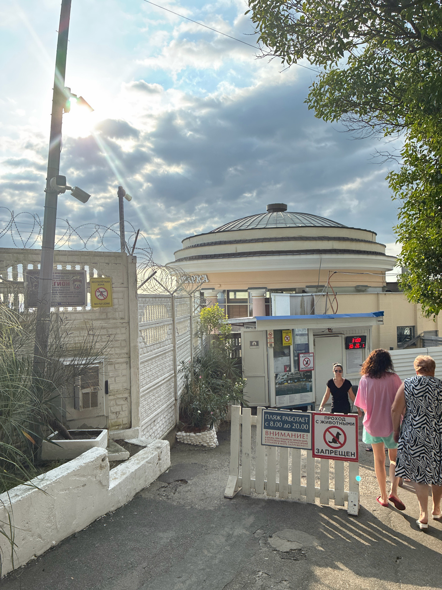 In Sochi, Russia, on a sunny day, three women walk past a white picket fence with several signs. The fence leads to a building with a large, circular, domed roof. One sign indicates that the beach is open from 8:00 to 20:00 and warns against entering the sea during a storm. Another sign prohibits passage with animals, depicting a dog with a red circle and slash over it. The women, dressed in casual summer clothing, are walking away from the camera. The environment appears to be an entrance to a public space, possibly a beach or recreational area, given the signage. The background shows a cloudy sky with bright sunbeams filtering through, and lush green trees.