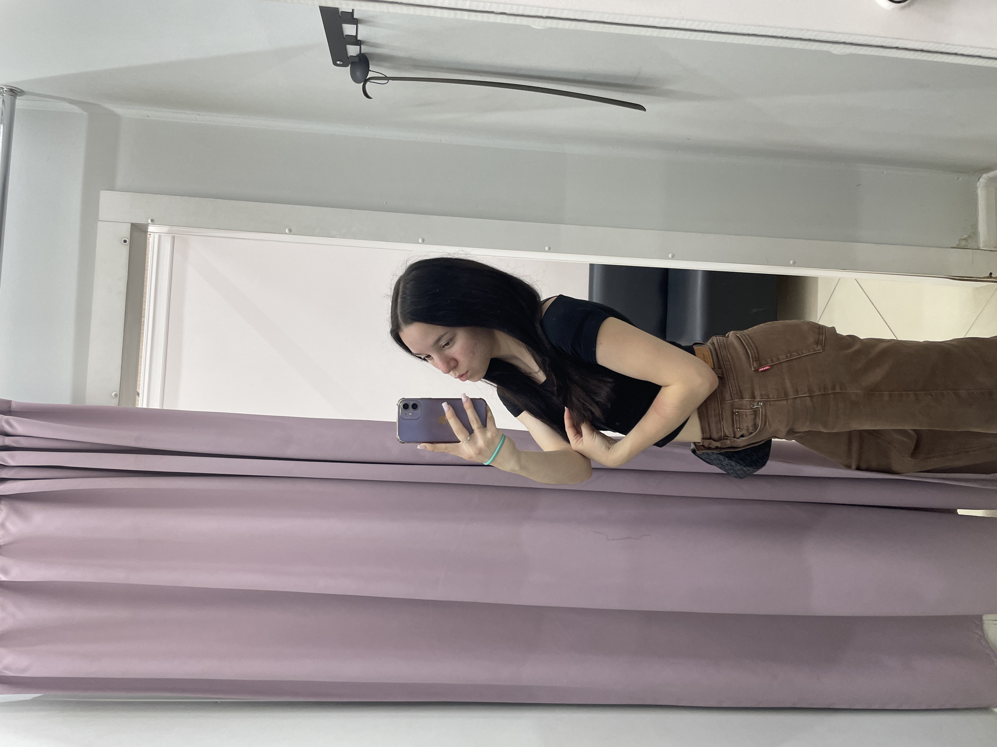 A young woman with long, dark hair is taking a mirror selfie in what appears to be a changing room. She is wearing a black crop top and brown pants. She is holding a purple smartphone in her right hand, angled to capture her reflection. A small, dark purse is slung across her body. The background consists of a light purple curtain on the left and a light grey wall on the right, with a black coat hook holding a dark, elongated object. The floor is tiled with a light color. The scene is indoors, likely in a retail environment.