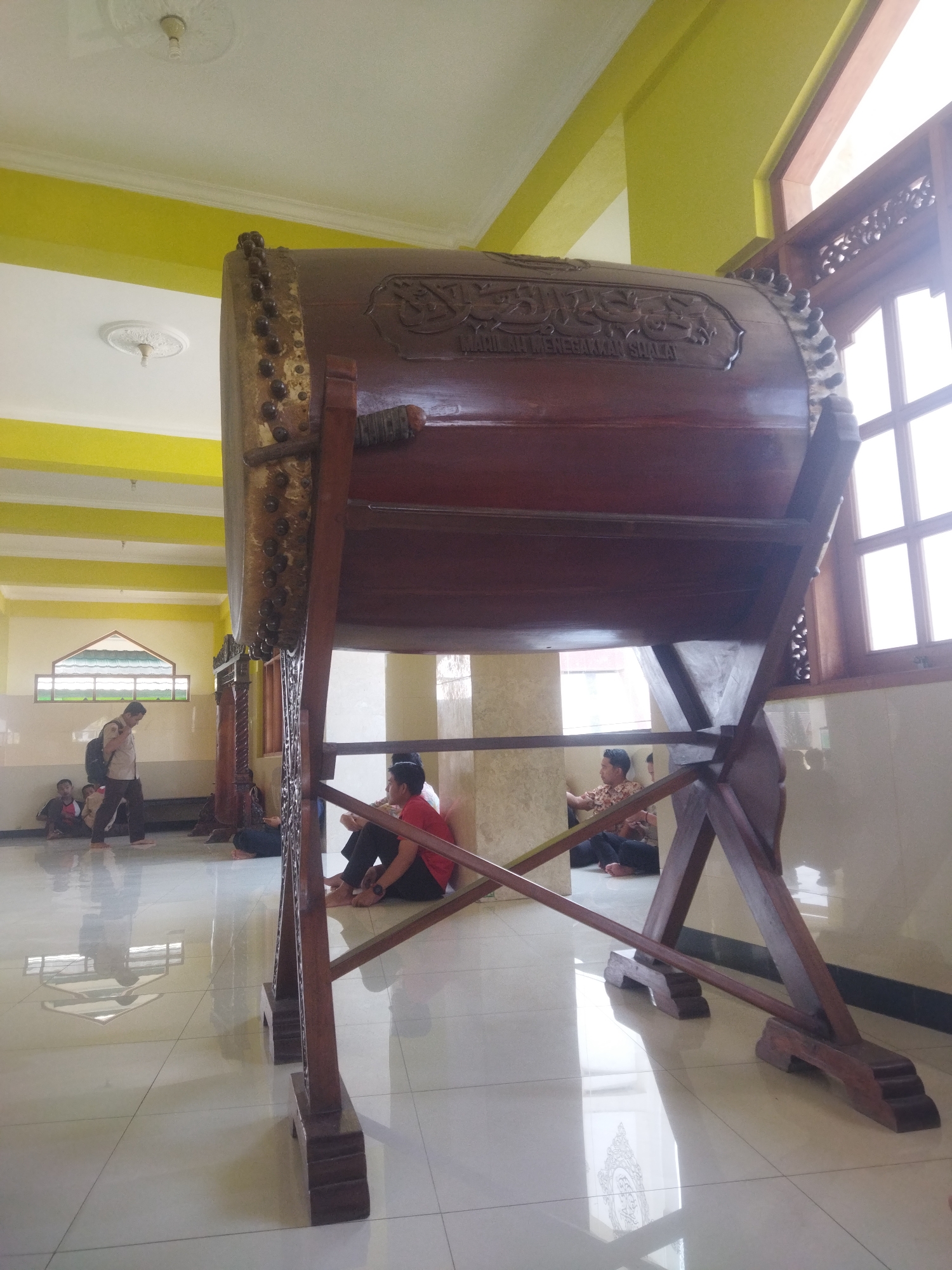 The image captures a large, ornate wooden drum, possibly a bedug, prominently displayed on a wooden stand within a building. The drum has intricate carvings and is adorned with studs along its circumference. Several individuals are present in the scene. In the foreground, to the right of the drum, a person in a red shirt and black pants is sitting on the floor, facing away from the camera. To the left, further in the background, a person in a light-colored shirt and dark pants with a backpack is walking, while several other individuals are seated on the floor nearby. The environment appears to be an indoor space with a tiled floor, yellow and white walls, and large windows that allow natural light to enter. The ceiling is white with decorative elements. The overall impression is that of a communal space, possibly a mosque or a community hall in Wonogiri, Indonesia, given the cultural context suggested by the drum.