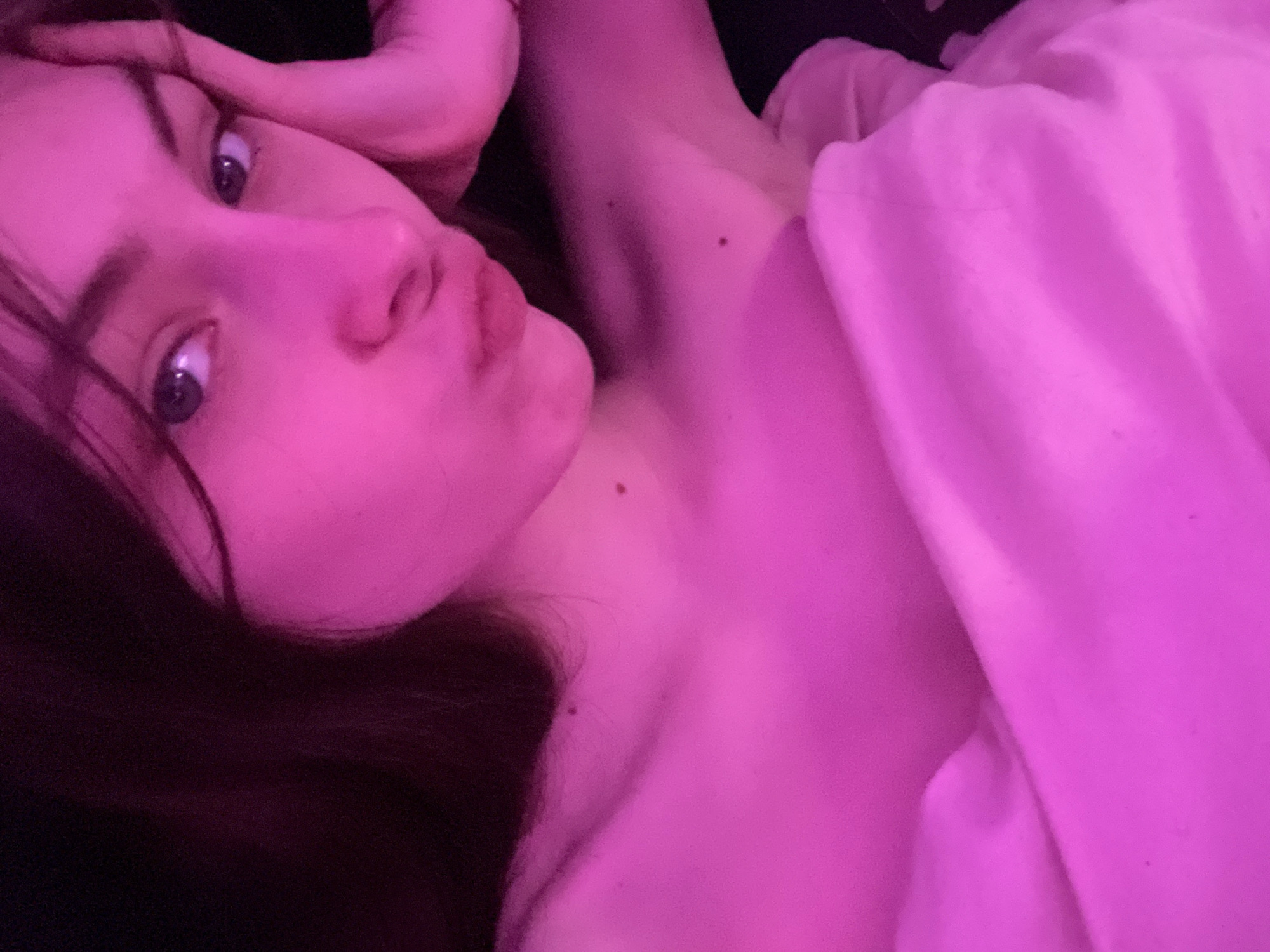 A young woman with dark hair and fair skin lies on a pink bed or under a pink blanket. Her head is tilted, and she is looking directly at the camera with her lips puckered in a kissing expression. One of her hands is near her forehead. The lighting is heavily saturated with pink, which affects the color of her skin and the surrounding environment. The overall scene appears to be indoors. There is no indication of the location being Saint Petersburg, Russia or any other specific city.