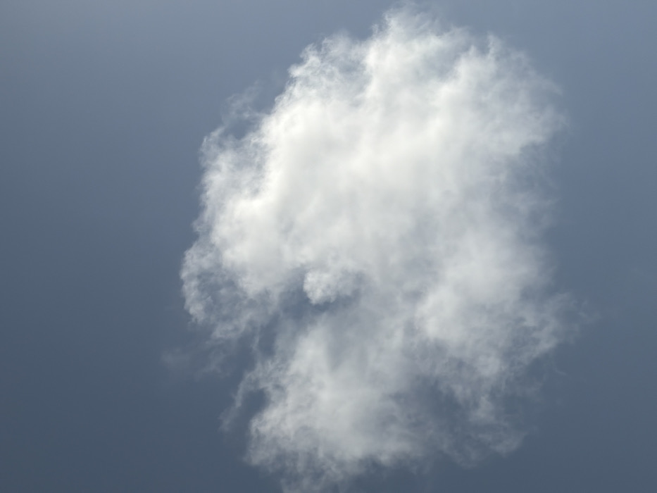 The scene depicts a singular, predominantly white cumulus cloud against a clear, uniform light blue-grey sky. The cloud, positioned roughly centrally within the frame, exhibits an irregular, somewhat rounded shape with diffuse, wispy edges extending from a denser core. Illumination suggests direct sunlight from an unseen source, highlighting portions of the cloud with brighter white tones, particularly towards the upper right. No persons, objects, or ground features are visible within the frame. The environment consists solely of atmospheric elements. The described location is Igboefon, Nigeria.