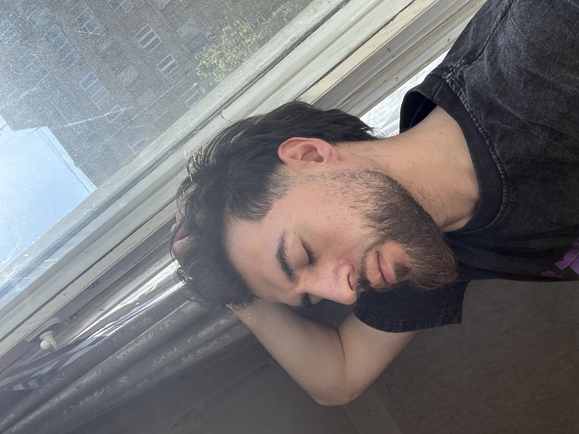 A young man with dark hair and a beard is depicted sleeping or resting peacefully next to an old window, his head cradled on his arm. His eyes are closed, and he wears a dark, possibly charcoal grey, t-shirt with a subtle purple design visible on the shoulder. The shot is angled, suggesting a perspective from above or to the side.
Through the window, which appears dusty or textured, an older, multi-story building with numerous windows is visible, indicating an urban environment. Bright sunlight streams in, suggesting it is daytime with a clear sky. Some green foliage is also visible outside, hinting at nearby trees or plants. The window frame itself is old and worn, painted in a light color, and features a visible white plastic latch and multiple panes. The general aesthetic of the building and window frame aligns with typical architecture found in cities like Yerevan, Armenia. No visible text is present in the image.