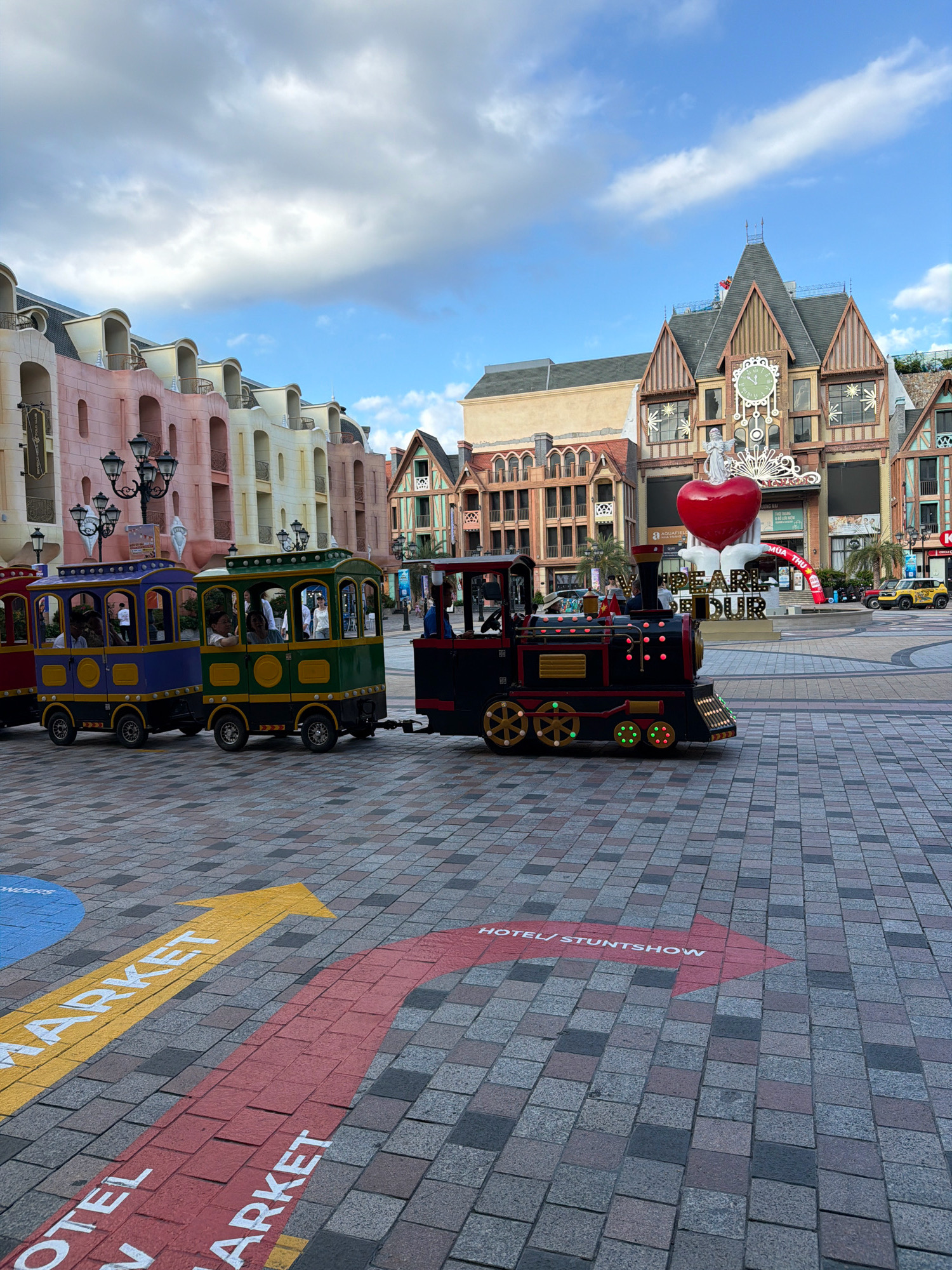 This is a full-frame, eye-level shot of a tourist train moving through a plaza in Nha Trang, Vietnam. The train, painted black with red and yellow accents, is comprised of a locomotive and three passenger cars, each a different color: the first is blue and yellow, the second green and yellow, and the third is partially visible and appears red. Several people, including adults and children, are seated in the passenger cars, looking in various directions. The plaza is paved with interlocking grey and reddish-brown bricks, some of which have painted arrows and text. One yellow arrow points left with "MARKET" written on it, a red arrow curves right with "HOTEL/STUNTSHOW" written on it, and a partial blue circle is visible to the left.
In the background, a cluster of European-inspired buildings line the plaza. The structures feature arched windows, decorative facades, and multi-pitched roofs. One prominent building in the center is a large, ornate structure with a clock face and a red heart sculpture at its entrance, supported by two hands. Palm trees are visible peeking out from behind some of the buildings. The sky above is partly cloudy with a mix of white clouds and blue sky. A yellow car is parked in the middle ground on the right. The lighting suggests it is daytime, possibly late afternoon, with soft shadows.