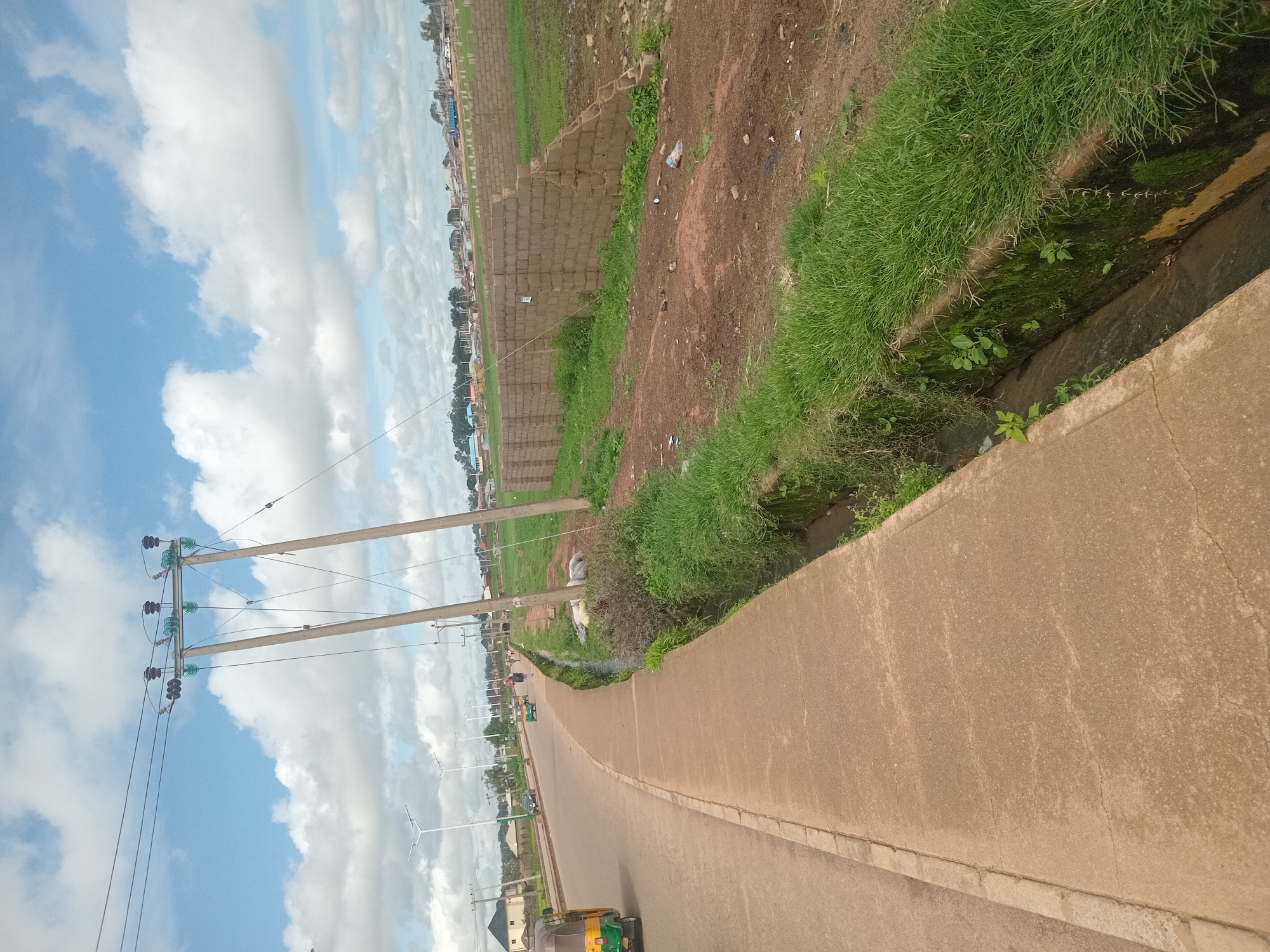 This image depicts a scene in Bukuru, Nigeria, likely an urban or semi-urban area. A paved road runs diagonally across the left side of the frame, with a concrete sidewalk bordering it. A three-wheeled vehicle, commonly known as an "okada" or "keke," is visible on the road. To the right of the sidewalk, there is a drainage ditch with overgrown green grass and moss on its concrete edges. Beyond the ditch, an undeveloped area is visible with a brick wall under construction, separating it from a grassy field. In the background, houses and other buildings are present, along with utility poles carrying power lines. The sky is cloudy with patches of blue. The overall impression is of a developing area with ongoing construction and typical elements of Nigerian urban infrastructure.