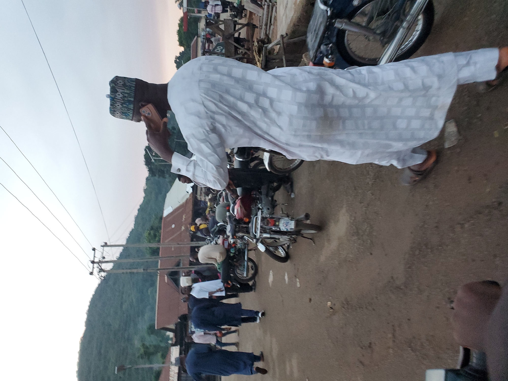 The image depicts an outdoor scene in Mararaba, Nigeria, characterized by human activity and local infrastructure. In the right-center foreground, a male individual is seen from behind, walking towards the right. He wears a long, loose-fitting white garment with a subtle grid pattern and a patterned cap (kufi) featuring blue, green, and white geometric designs. He is holding a gold-colored smartphone to his right ear, engaged in a conversation. Brown sandals are visible on his feet.
The mid-ground is populated by multiple individuals and motorcycles. To the left, two men dressed in dark blue traditional Nigerian attire (agbada) walk on an unpaved road. Other individuals in varying attire are scattered throughout, some standing, others seated on motorcycles. A man in a light-colored, possibly beige, long-sleeved shirt is seated on a dark motorcycle in the center-right mid-ground. Numerous motorcycles, consistent with models commonly used for transportation in Nigeria, are parked or standing on the unpaved ground, which appears to be packed dirt or gravel. One motorcycle in the far-right foreground shows a silver exhaust pipe. A motorcycle further back displays a partially visible white license plate.
In the background, multiple single-story buildings with reddish-brown corrugated metal roofs are visible, suggesting a residential or market area. Utility poles with overhead electrical wires traverse the upper mid-ground. The distant background features a range of green, tree-covered mountains under a light, overcast sky, indicative of either early morning or late afternoon lighting.