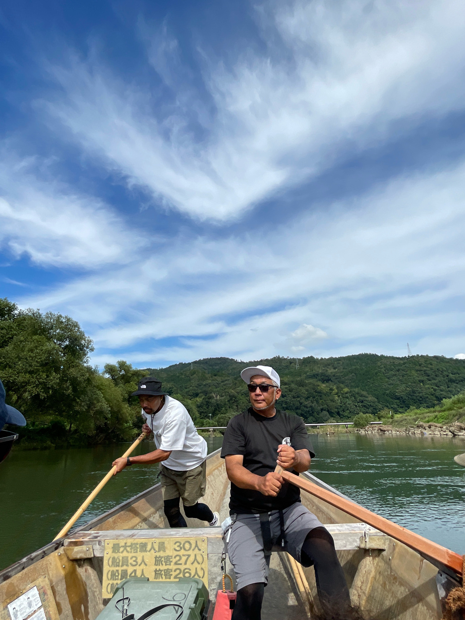 This is a medium shot, eye-level, outdoor, vertical photograph taken on a sunny day. Two adult males are in a wooden boat on a river, rowing. The boat is weathered and yellow, with signs on its interior side. The man in the foreground is on the right, wearing a black t-shirt, grey shorts, a white baseball cap, and sunglasses. He is actively rowing with a wooden oar. The man in the background is on the left, wearing a white t-shirt, khaki shorts, a black bucket hat, and boots. He is also rowing with a wooden oar. The river water is green and appears calm. Lush green trees and a forested hill are visible on the riverbanks. The sky is blue with scattered white, wispy clouds. The scene is likely the Hozugawa River in Kameoka, Japan, known for its scenic boat rides.