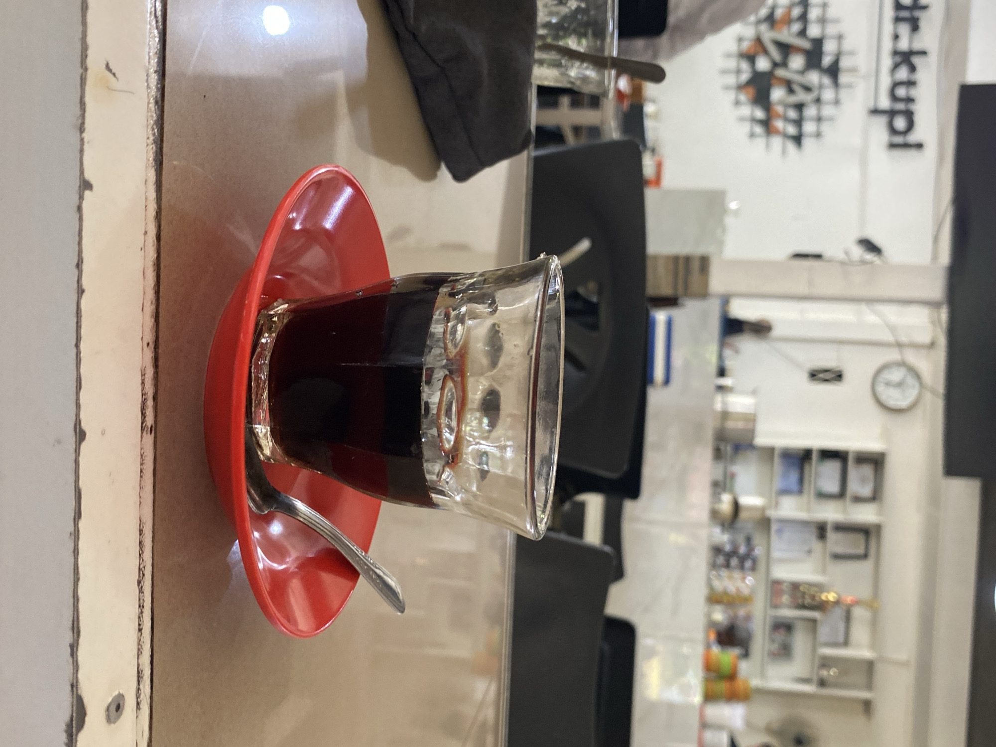 This is a medium shot of a glass of dark liquid, likely coffee, sitting on a red saucer with a silver spoon. The glass has a faceted design and contains a dark liquid with some reflection on its surface. The red saucer is glossy and reflects light. The scene appears to be set on a light brown, textured table. In the background, out of focus, are tables, chairs, and shelving units which suggest an indoor setting like a cafe or restaurant. On the wall in the background, the text "dr. kupl" is visible in large black lettering. There is also a circular graphic with black and orange elements. Further back, shelves hold various items, including what appear to be framed certificates and decorative objects, and a wall clock. The overall lighting is somewhat dim. The location of the establishment "dr. kupl" is Lhokseumawe, Indonesia, as indicated by the brand name which is a common coffee shop chain in that region.