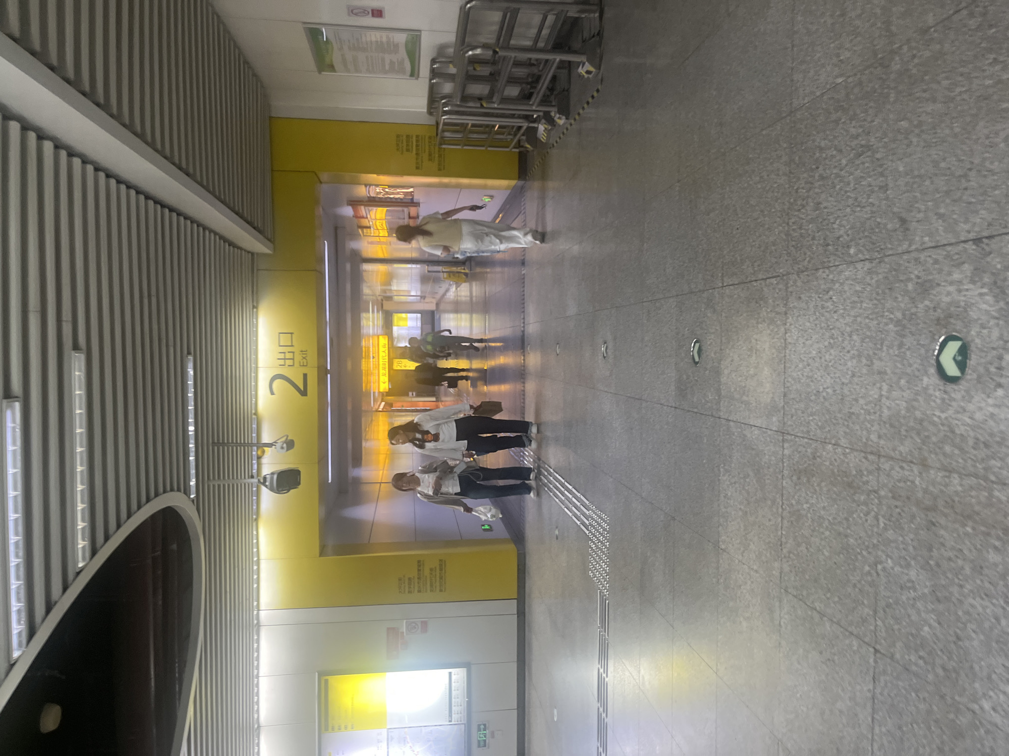 The image depicts an indoor scene, likely a subway station or a large public transit hub, in Yuzhong District, China. The environment is characterized by a modern, clean aesthetic with polished gray tiled floors and a suspended ceiling with integrated lighting. A prominent yellow-painted wall section forms an archway in the background, with the number "2" and the word "Exit" in both Chinese and English displayed above it.
In the foreground, a tactile paving strip is visible on the floor, indicating accessibility features. Several people are present, walking through the space. Two women are walking towards the viewer's left, in front of the yellow archway. One is wearing a white top and dark pants, and the other has on a white top, light cardigan, and dark pants. Further back, through the archway, other individuals are seen walking away from the camera, including a person in light-colored clothing walking in the center and a person in darker attire to the left.
The overall impression is of a well-lit, functional public space with clear signage and pedestrian flow. Barriers, possibly for crowd control or directing traffic, are visible on the right side of the frame. The lighting appears to be a mix of overhead fluorescent and recessed lighting, casting a bright illumination on the scene.