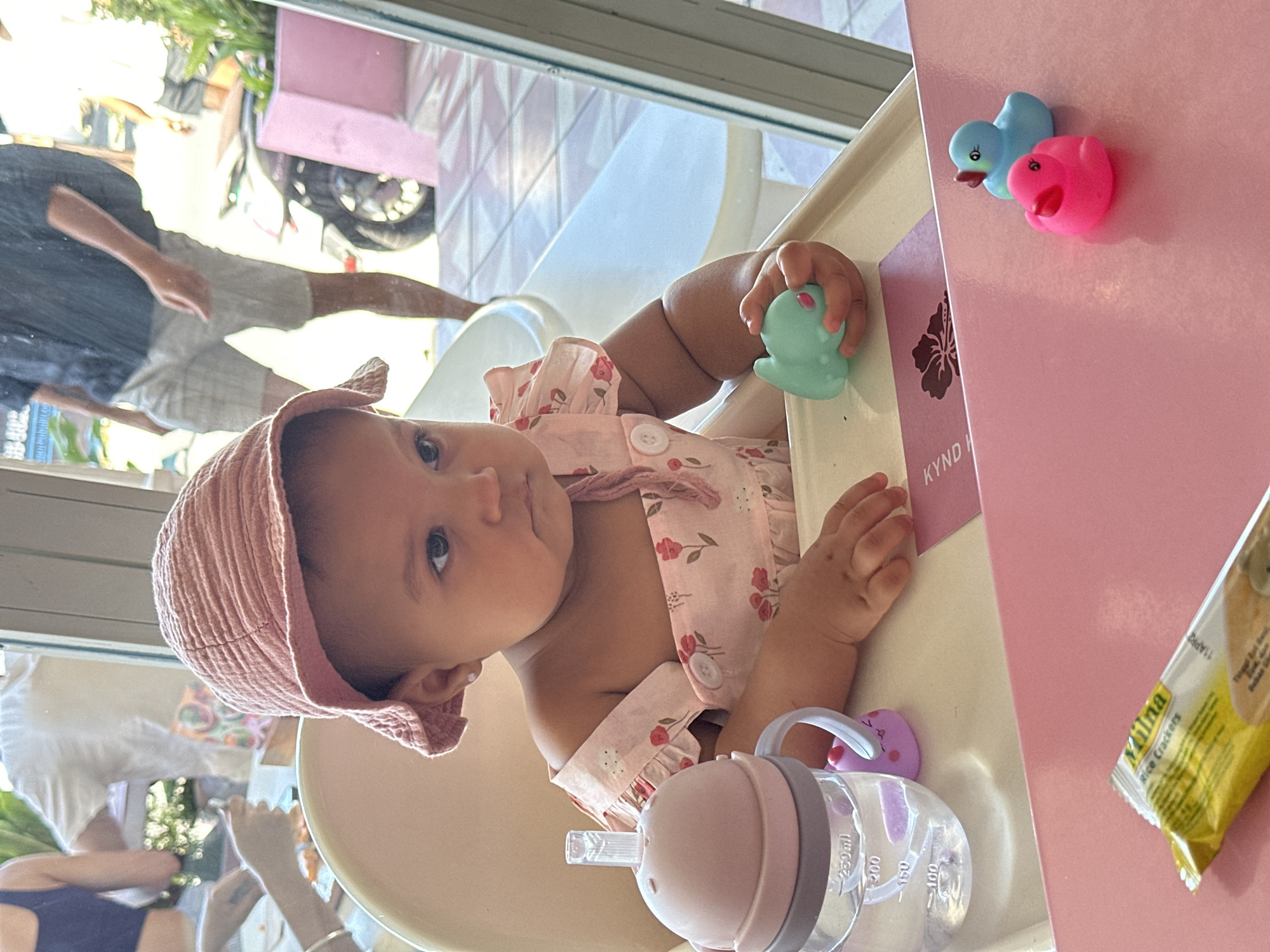 A baby wearing a pink hat and a floral-patterned outfit is sitting in a high chair. The baby is holding a green toy. On the high chair tray, there is a pink bottle with markings for measuring volume, and a pack of Milna rice crackers. A blue and pink duck are stuck to the side of the high chair, which also has a pink card with the word "KYND" printed on it. Through the window, one can see a reflection of people and a motorbike. The location is identified as North Kuta, Indonesia, based on the style of the building.
